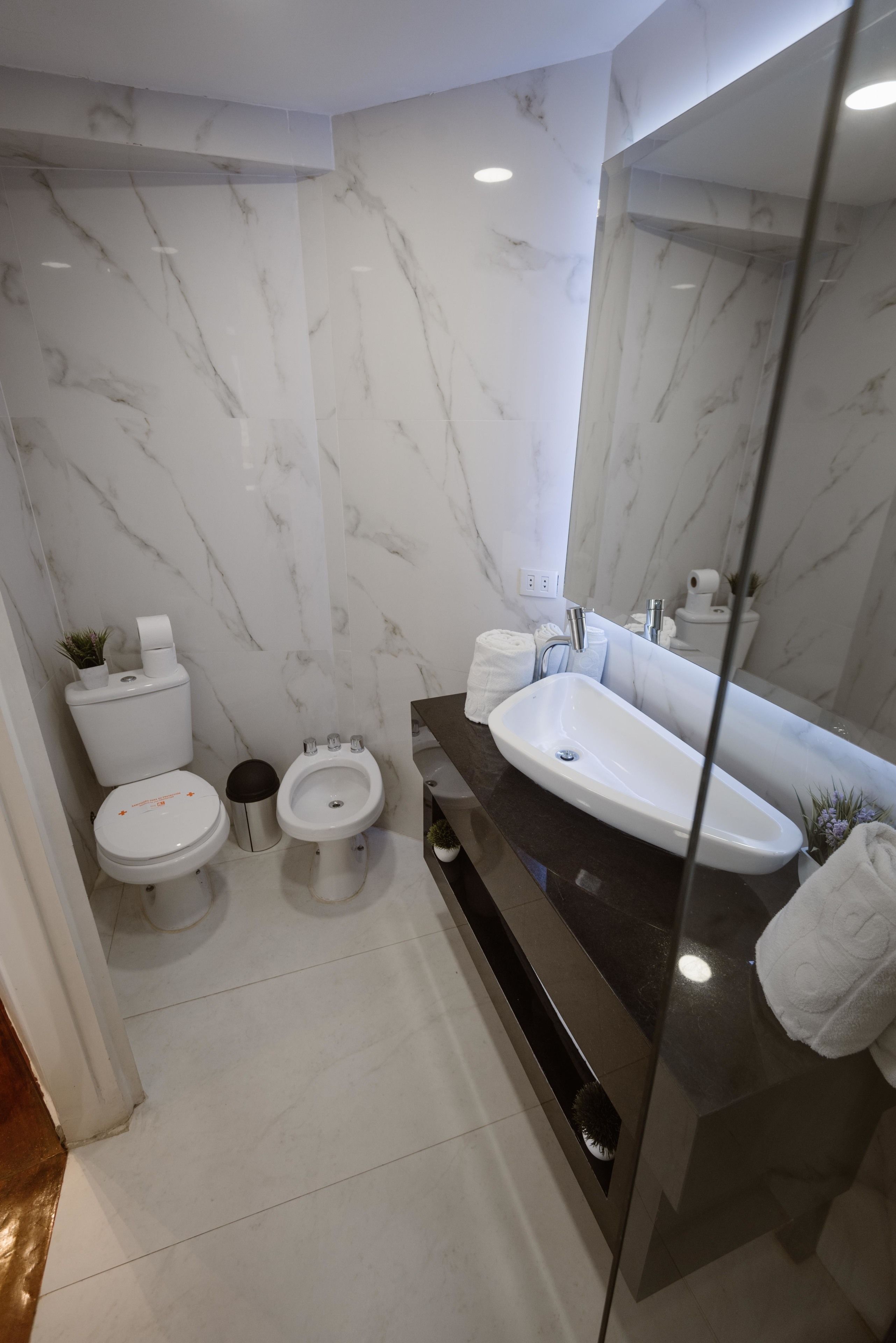 Un baño moderno con pared de mármol, encimera negra, lavamanos blanco y dos inodoros.