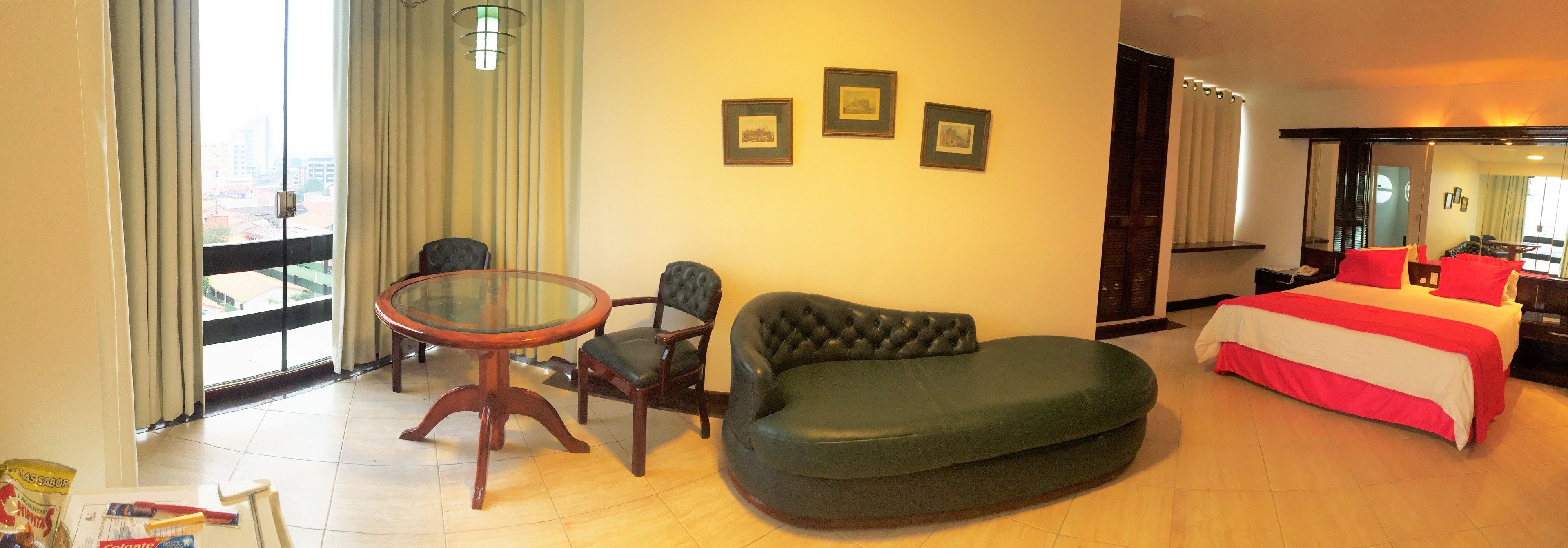A hotel room with a bed, a green leather sofa, a glass-top table, two chairs, and a large window with curtains.