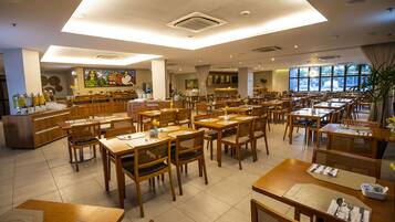 A spacious dining area with multiple wooden tables and chairs, a central buffet station, and large windows providing natural light.