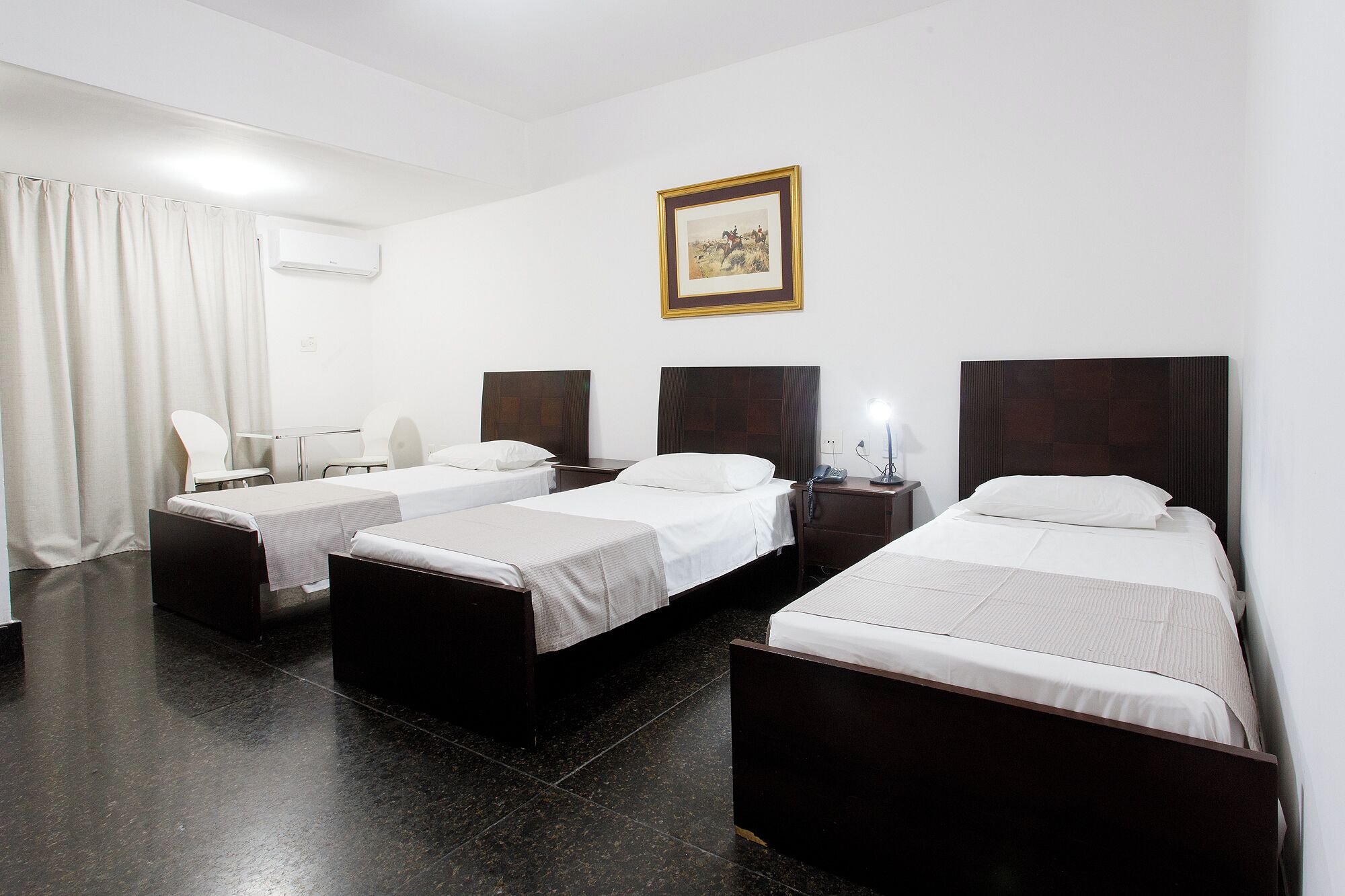 A hotel room with three single beds, a desk with a chair, a bedside table with a lamp, and a framed picture on the wall.