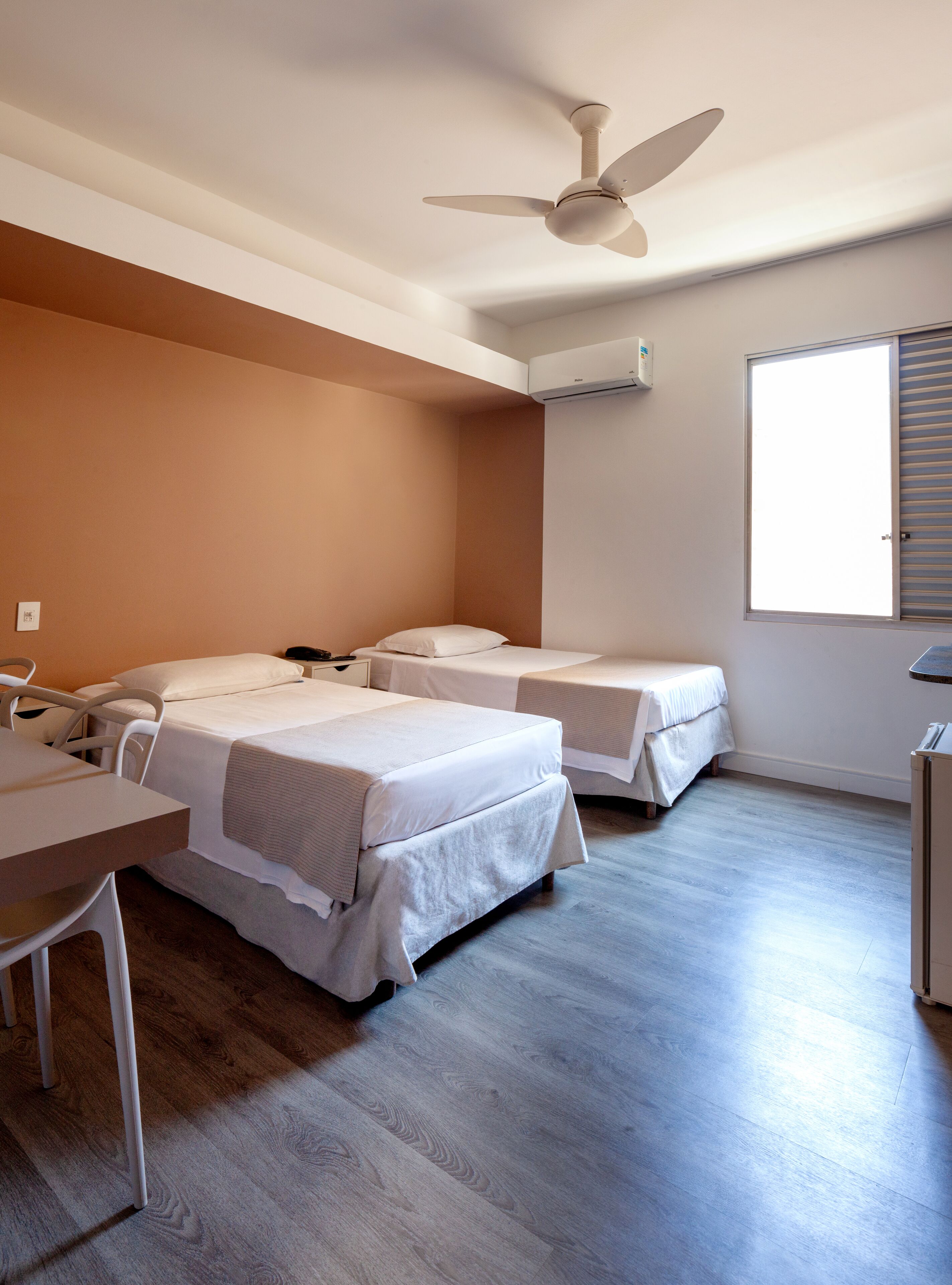 Habitación de hotel con dos camas, un escritorio, una silla, ventilador de techo, unidad de aire acondicionado y ventana con persianas.