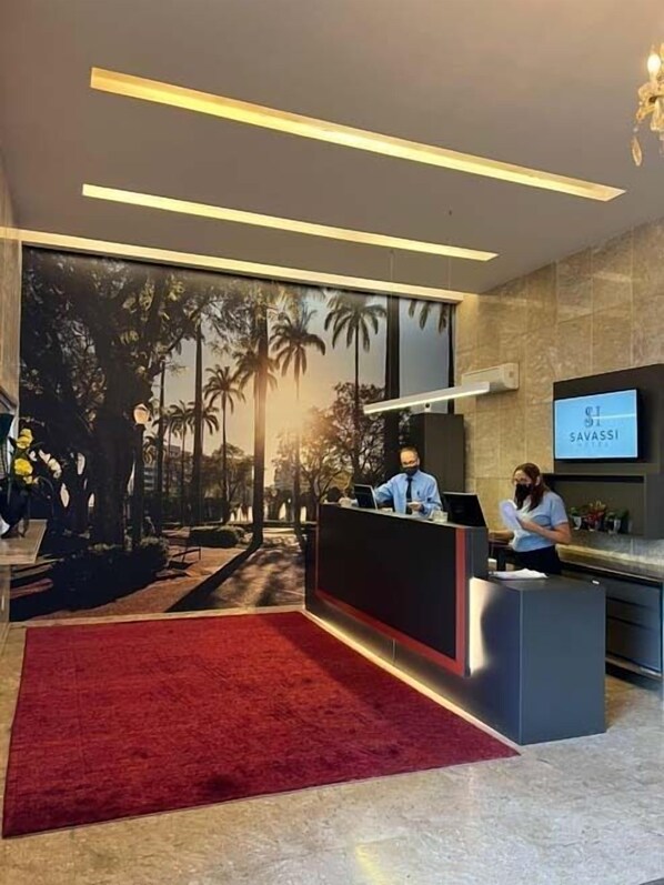 Un espace d’accueil avec un tapis rouge, une grande fenêtre donnant sur un paysage tropical et une enseigne indiquant « SAVASSI ».