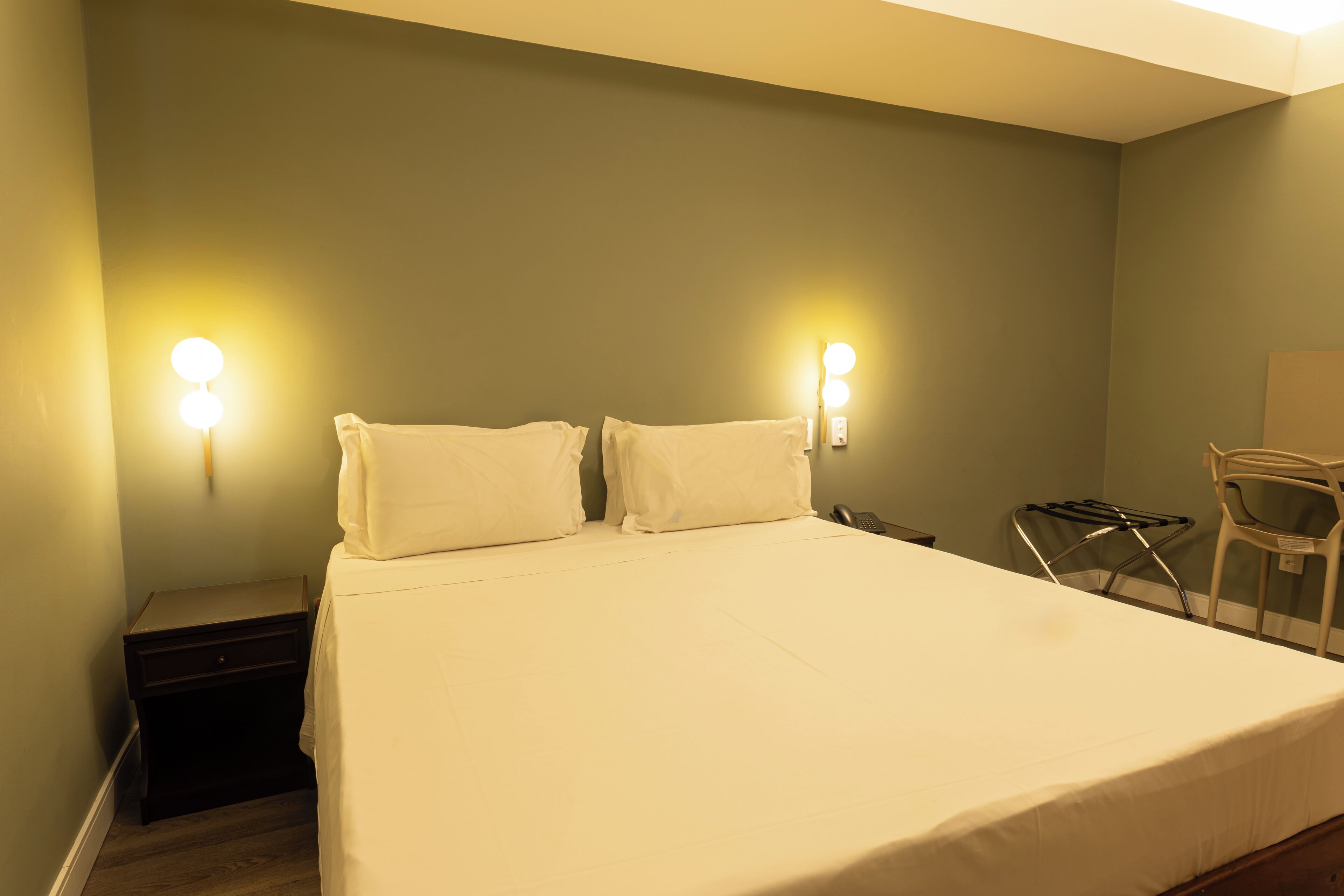 Una cama bien hecha con sábanas blancas en una habitación con paredes verdes y dos lámparas fijadas a la pared.