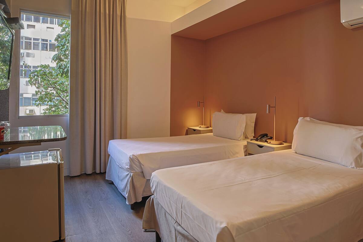 Une chambre d’hôtel avec deux lits, un bureau, une chaise, une petite table, une table de chevet et une fenêtre avec des rideaux.
