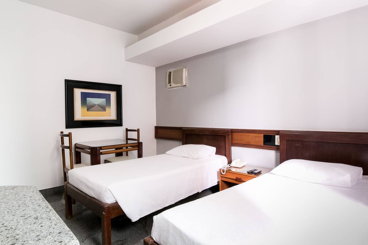 Une chambre d’hôtel avec deux lits, un bureau, une chaise, un tableau encadré, un téléphone et une bouche d’aération fixée au mur.