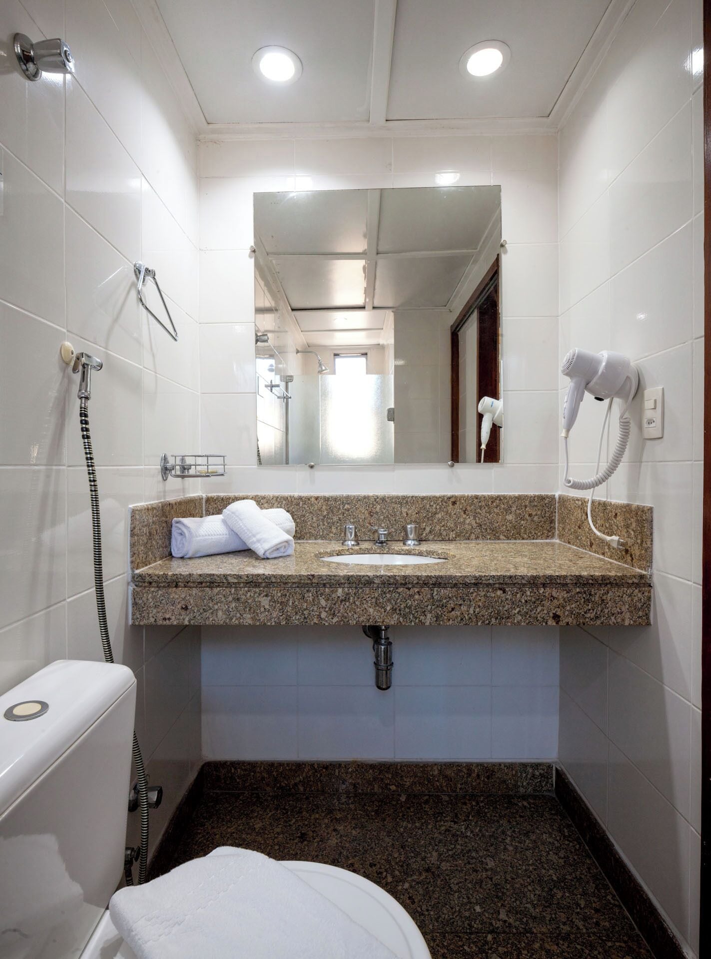 Un baño con encimera de granito, un lavabo, un espejo, un perchero para toallas, un secador de pelo y una ducha.