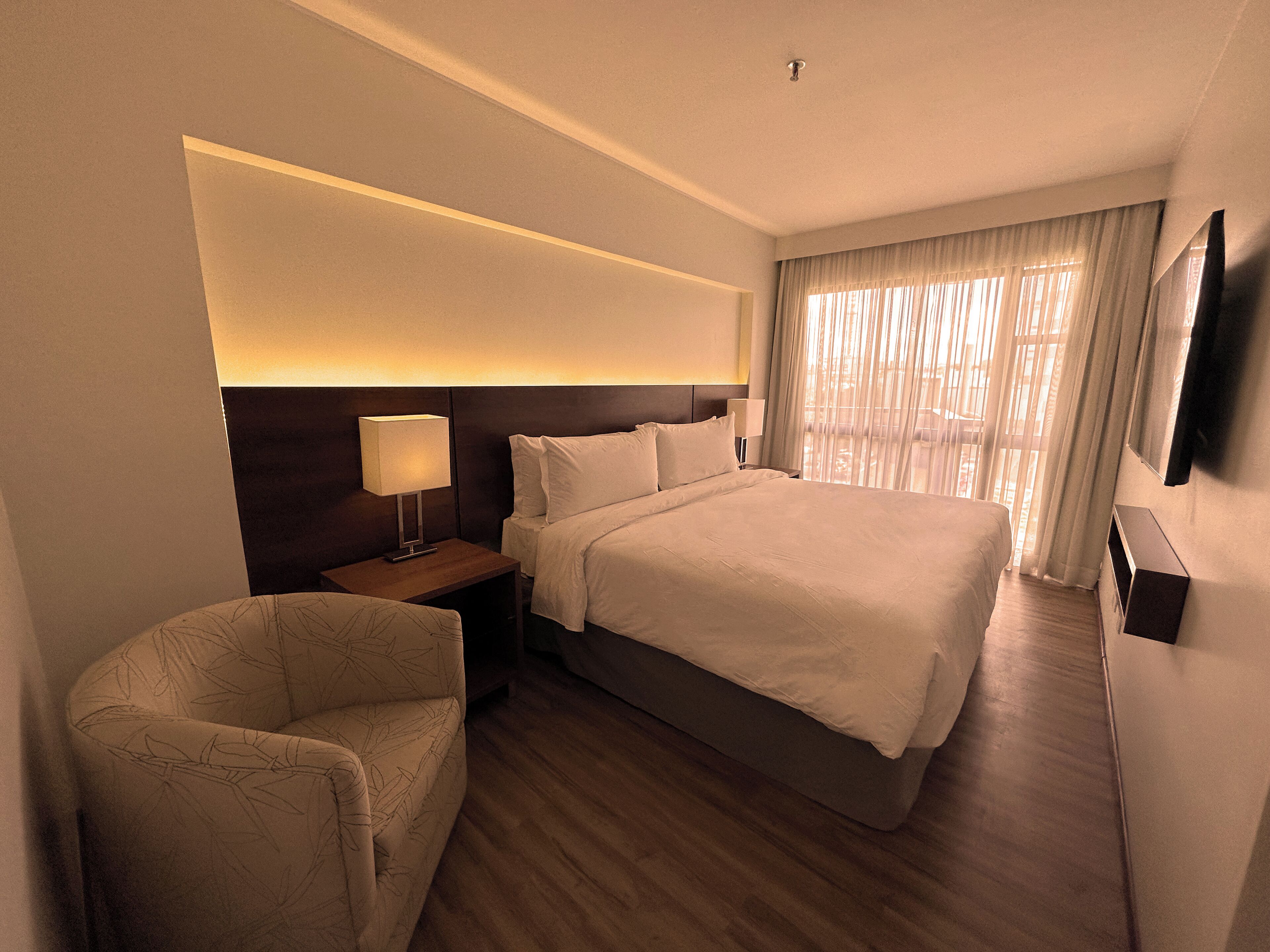 A hotel room with a large bed, a bedside table with a lamp, a chair, and a TV mounted on the wall.