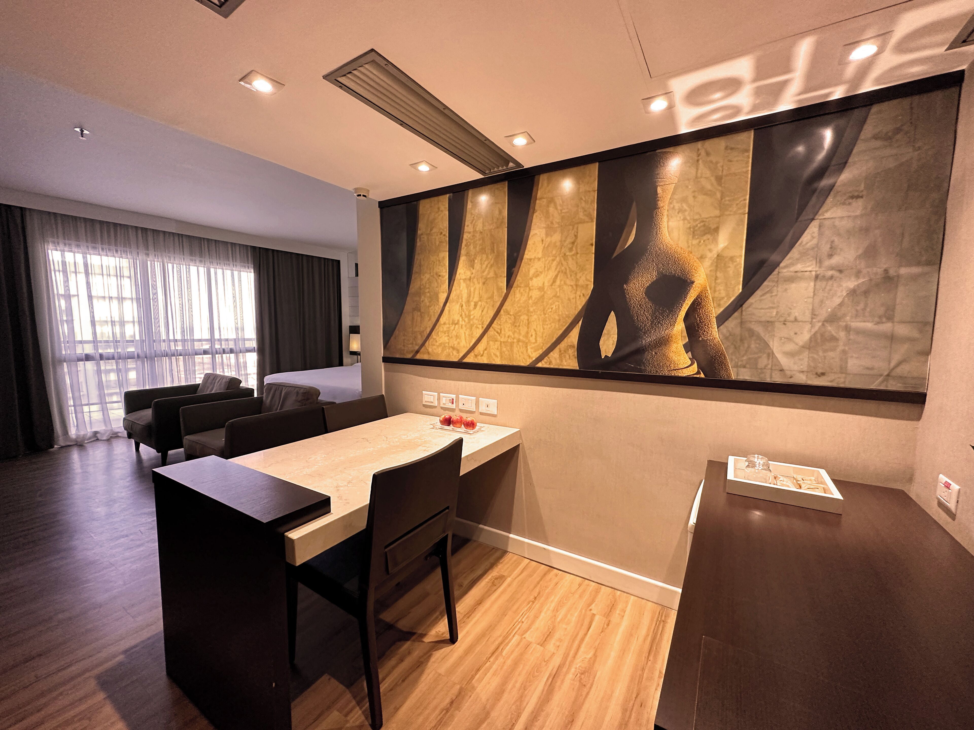 Quarto de hotel moderno com uma cama grande, área de refeições com mesa e cadeiras, um grande quadro abstrato na parede e vista da cidade pela janela.