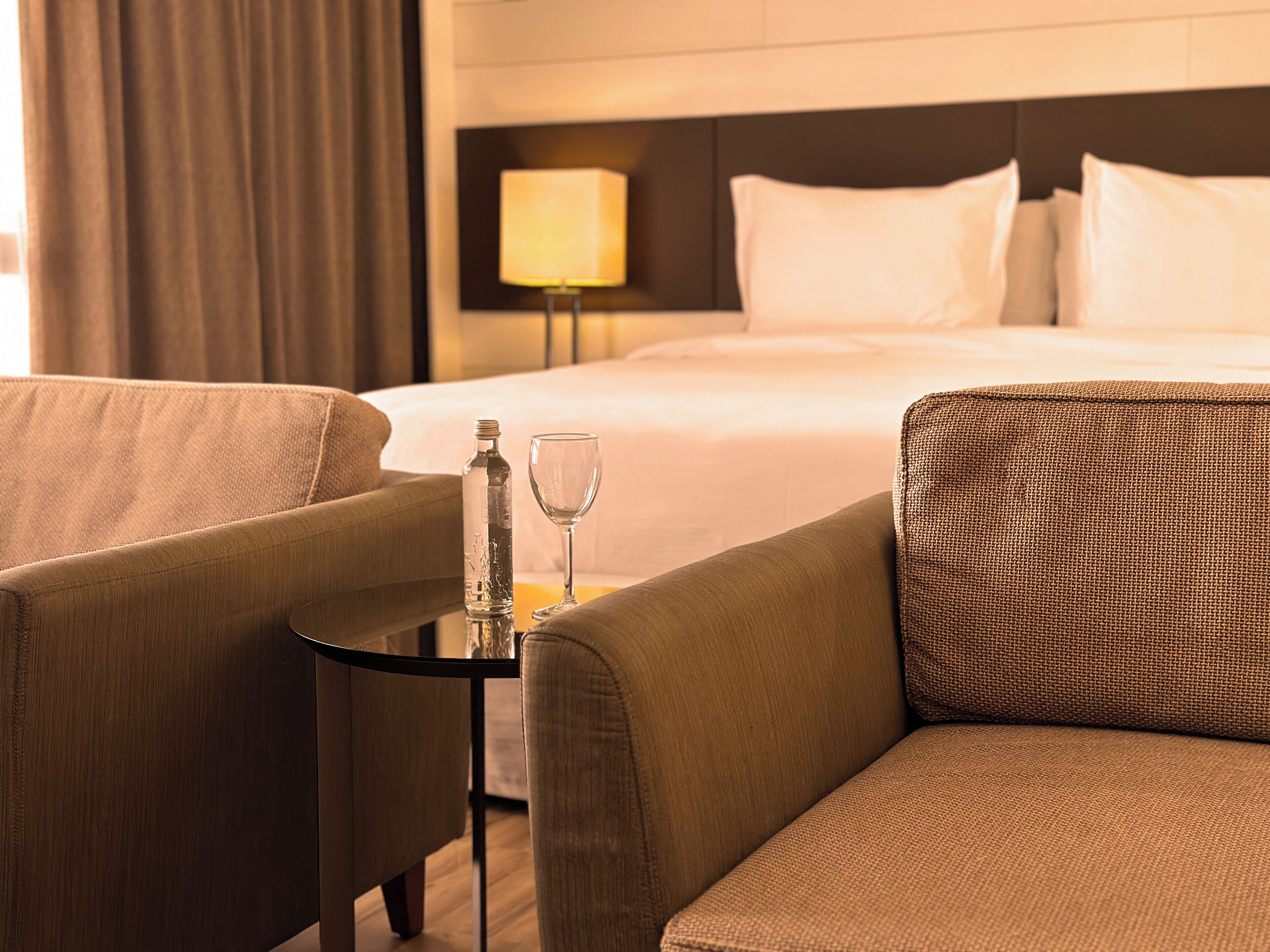 Ein Hotelzimmer mit einem Bett, zwei Sesseln, einem kleinen Tisch mit Glas und Flasche sowie einem Nachttisch mit Lampe.