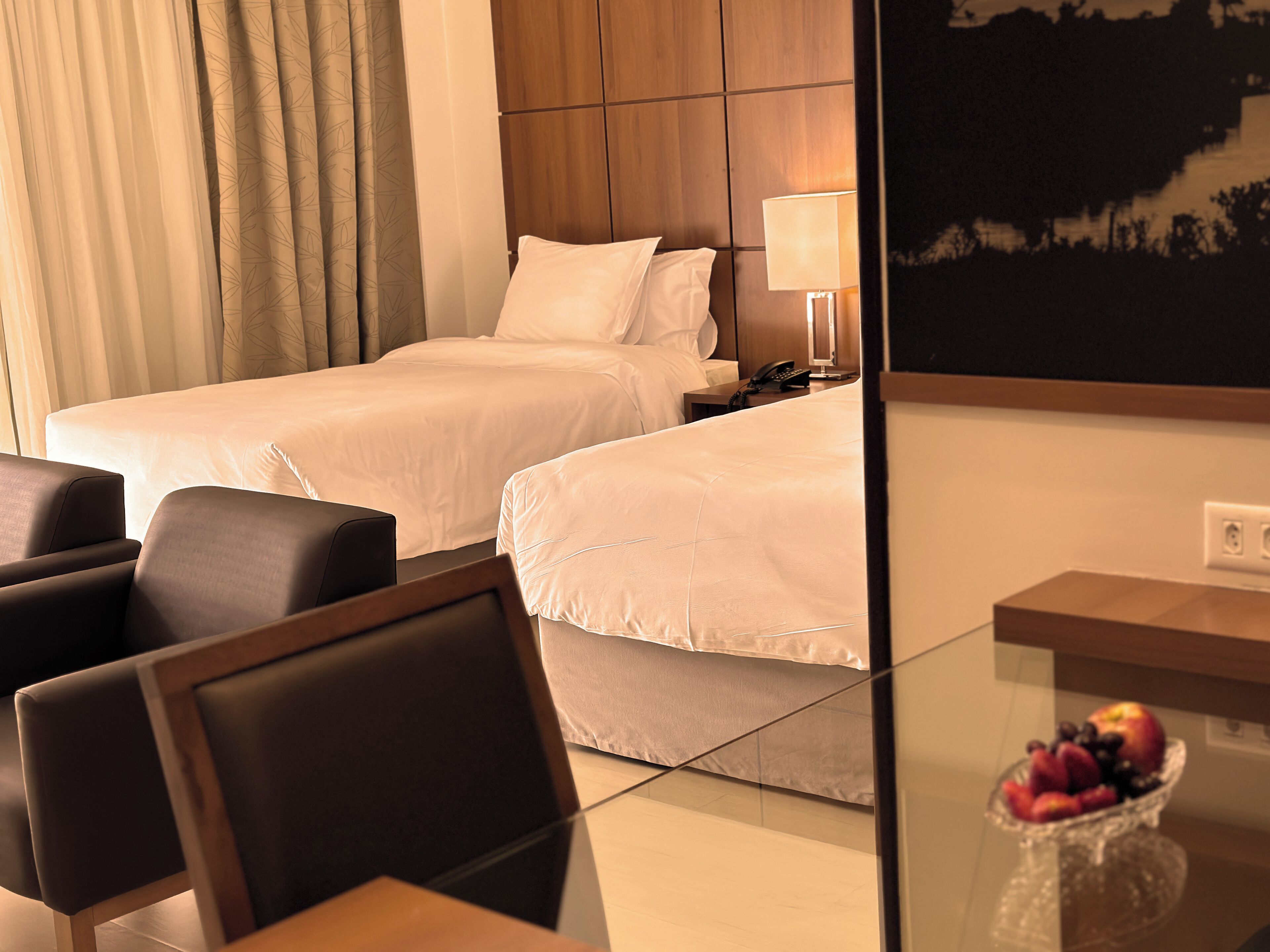 A hotel room with two beds, a desk with a lamp, a chair, and a fruit bowl on the desk.