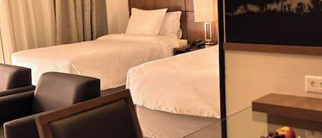 A hotel room with two beds, a desk with a lamp, a chair, and a fruit bowl on the desk.
