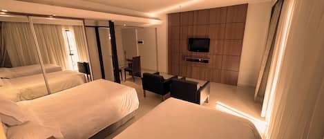 Habitación de hotel con dos camas, un televisor montado en la pared, una zona de estar y una mesa de comedor.