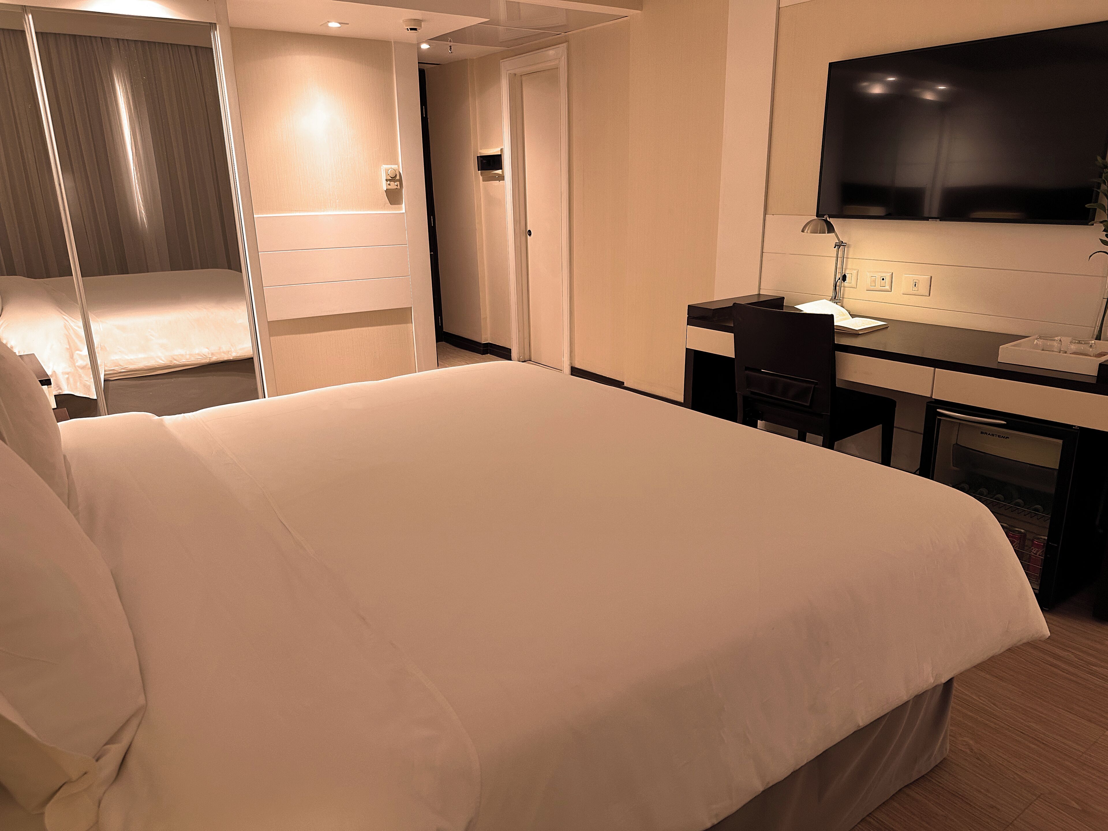 Ett hotellrum med en stor säng, ett skrivbord, en stol, en platt-TV och en minibar.
