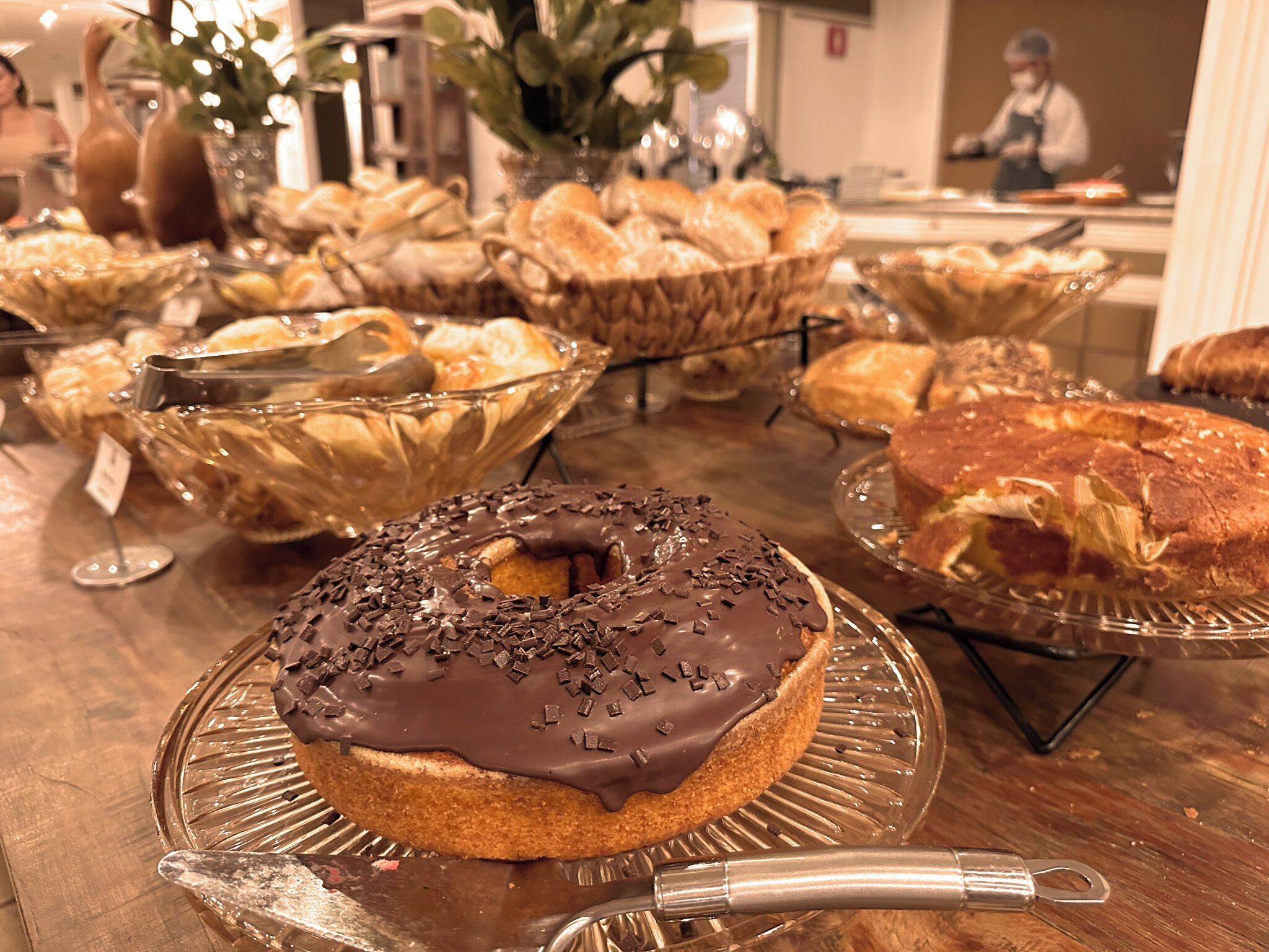Diversas opciones de productos horneados expuestos sobre una mesa de madera, incluyendo un donut con glaseado de chocolate, cestas de pan y pastelería.