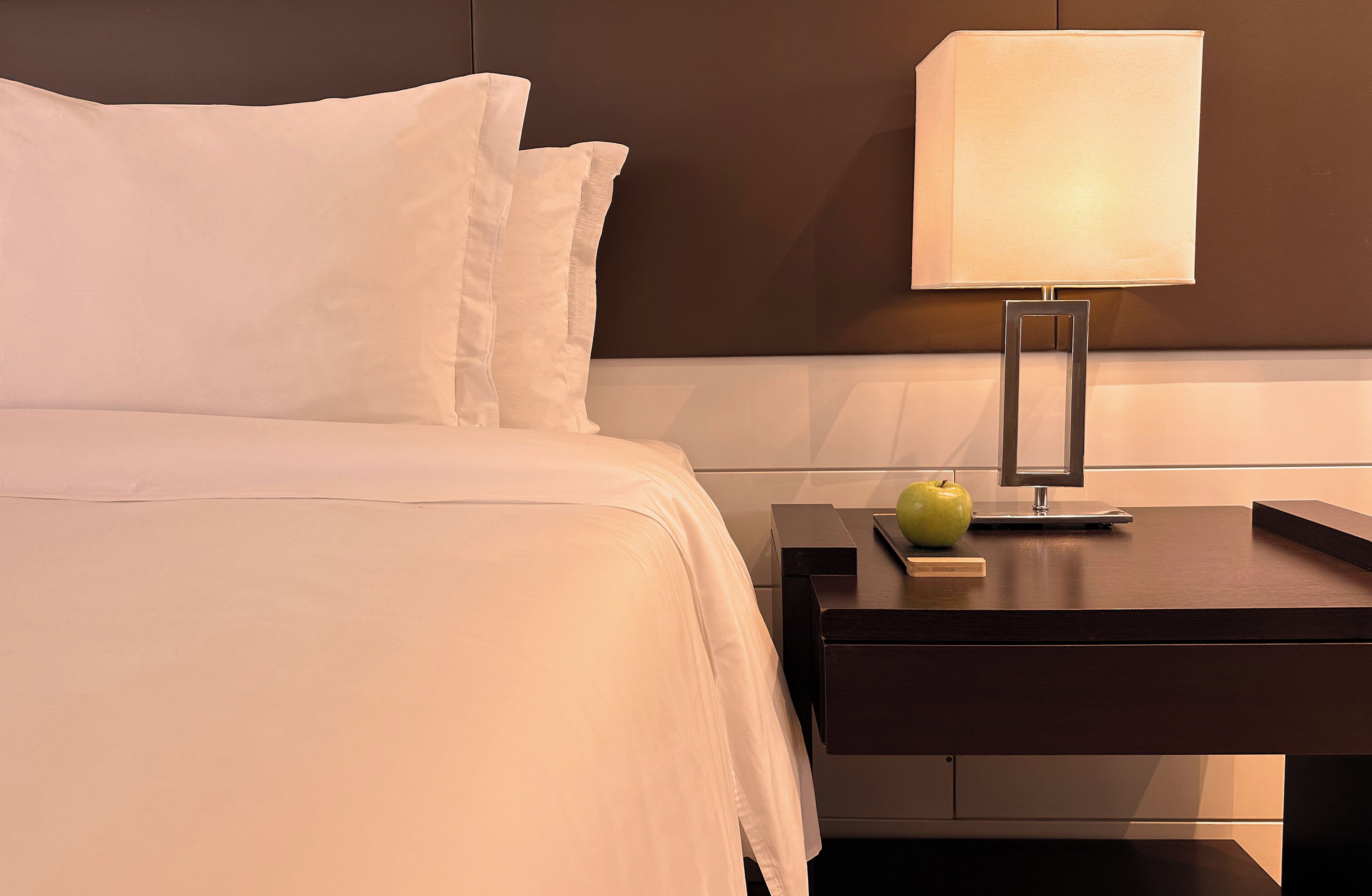 A neatly made bed with white linens, a bedside table with a lamp, and a green apple on the table.