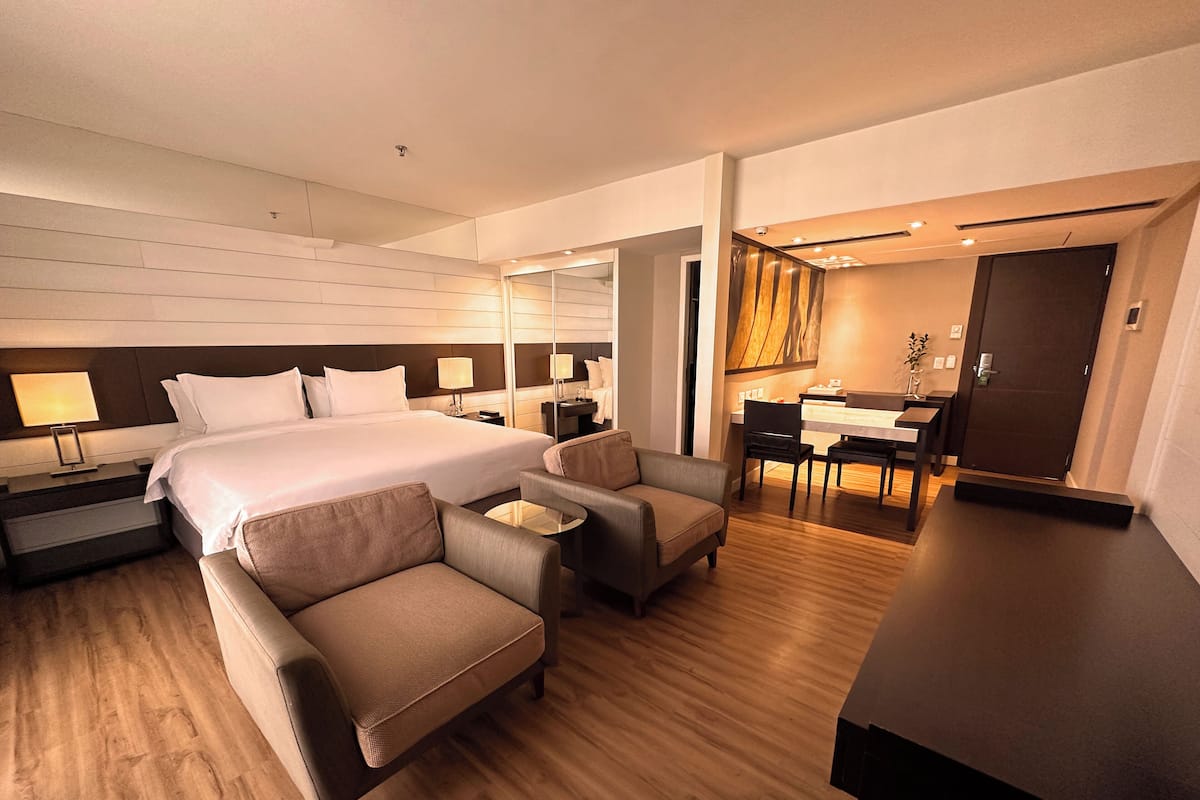 Una habitación de hotel moderna con una cama grande, un área de descanso con dos sillones, una mesita, un escritorio con una silla y un espejo.