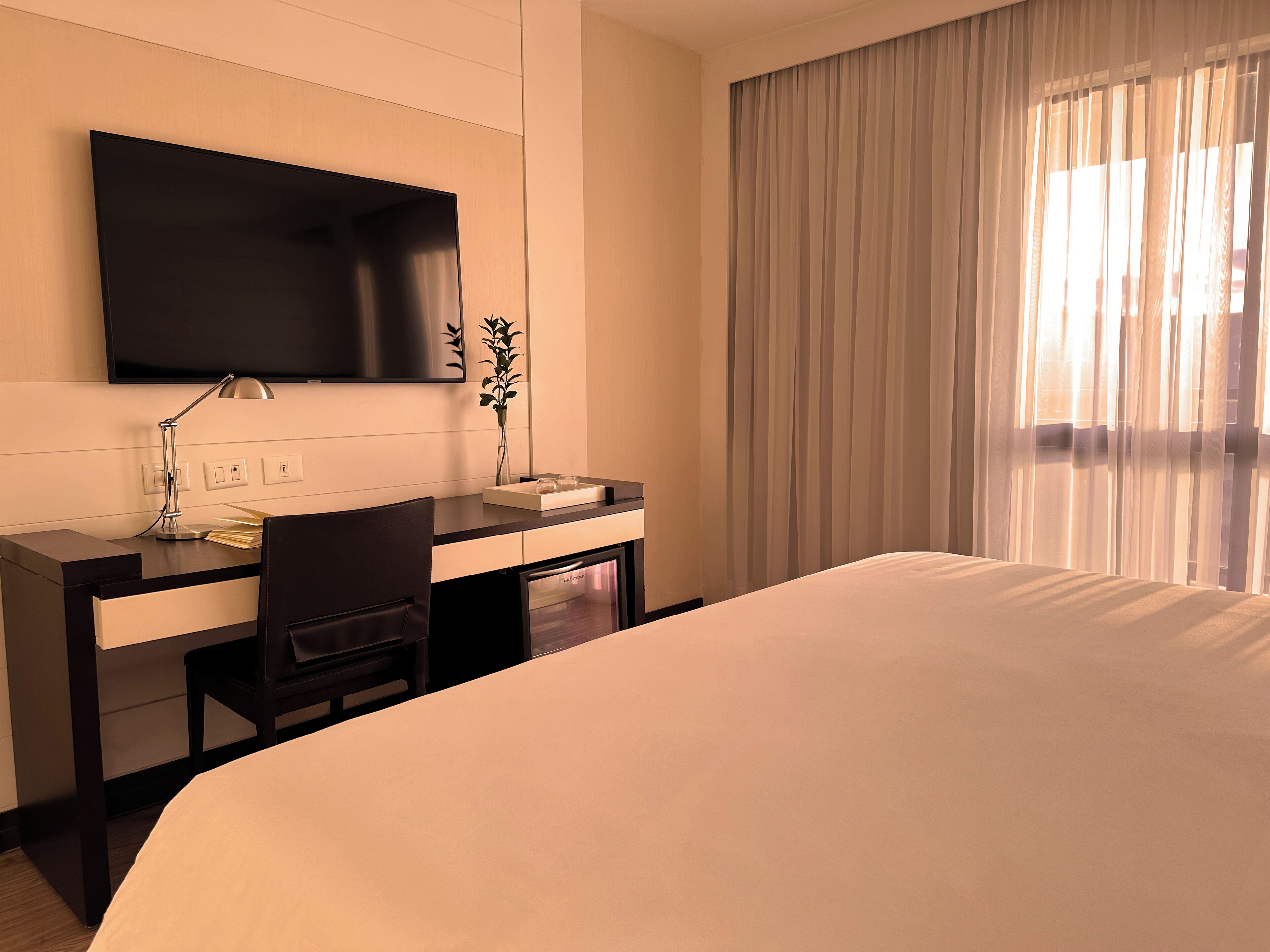 Quarto de hotel com cama, escrivaninha com cadeira, televisão na parede e janela com cortinas.