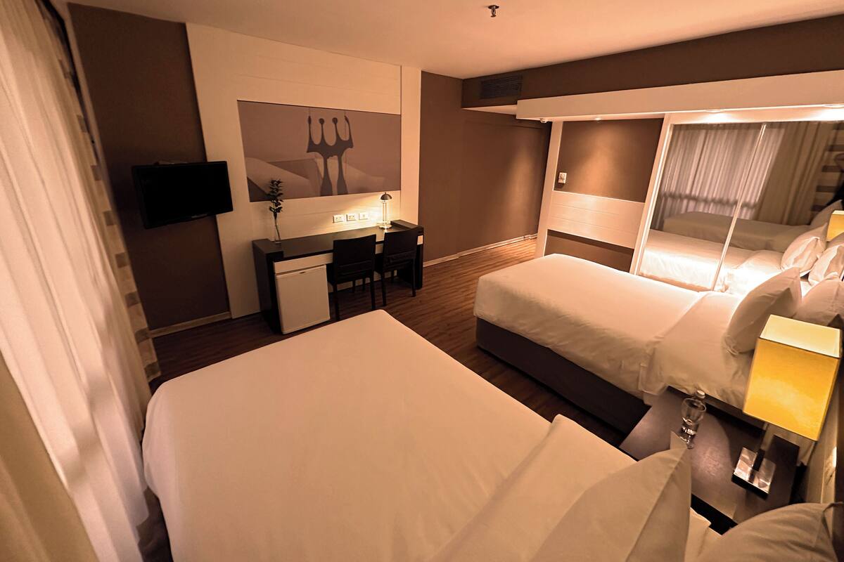 Habitación de hotel con cama, escritorio con silla, refrigerador pequeño, televisor montado en la pared y un espejo grande.
