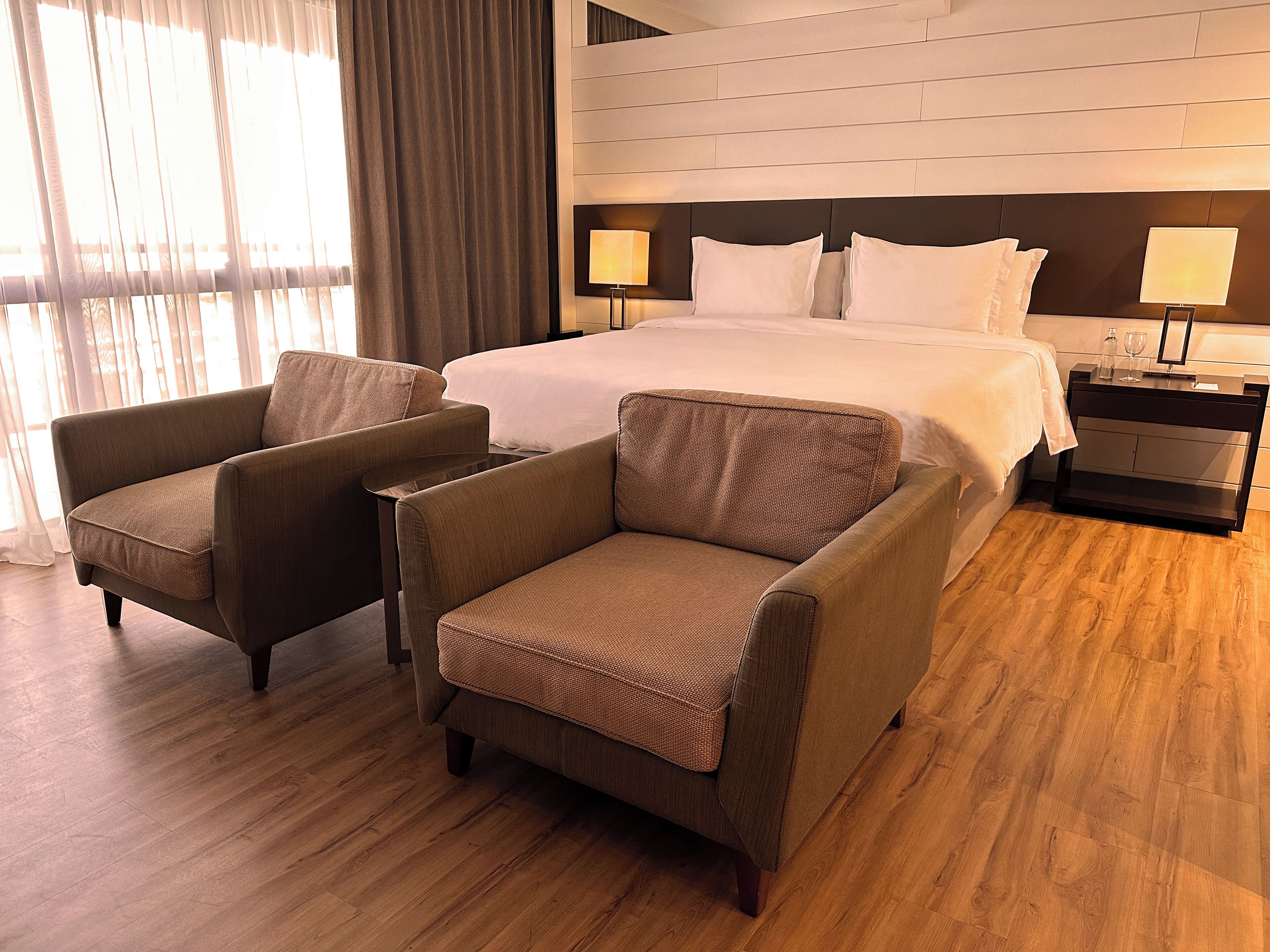 Une chambre d’hôtel avec un grand lit, deux fauteuils, une table de chevet avec une lampe et un plancher en bois.
