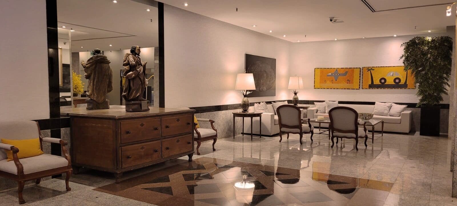 A modern hotel lobby with a marble floor, a wooden dresser, a sofa, and a coffee table. There is a large mirror on one wall and artwork above the sofa.