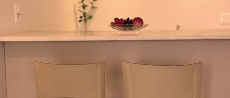 A kitchen with a white countertop, two beige chairs, a vase with green plants, and a bowl of fruit.
