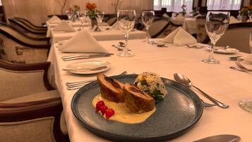 A dining table set with plates, cutlery, and glasses, with a dish of food in the foreground.