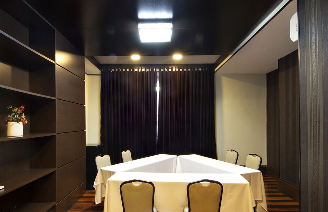 A conference room with a rectangular table, chairs, a white tablecloth, a shelf with a vase of flowers, and a dark curtain backdrop.