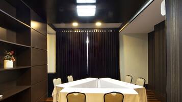 A conference room with a rectangular table, chairs, a white tablecloth, a shelf with a vase of flowers, and a dark curtain backdrop.