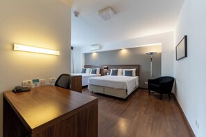 A hotel room with a wooden desk, two beds, a chair, and a standing lamp. - Hotel Deville Curitiba (Curitiba)