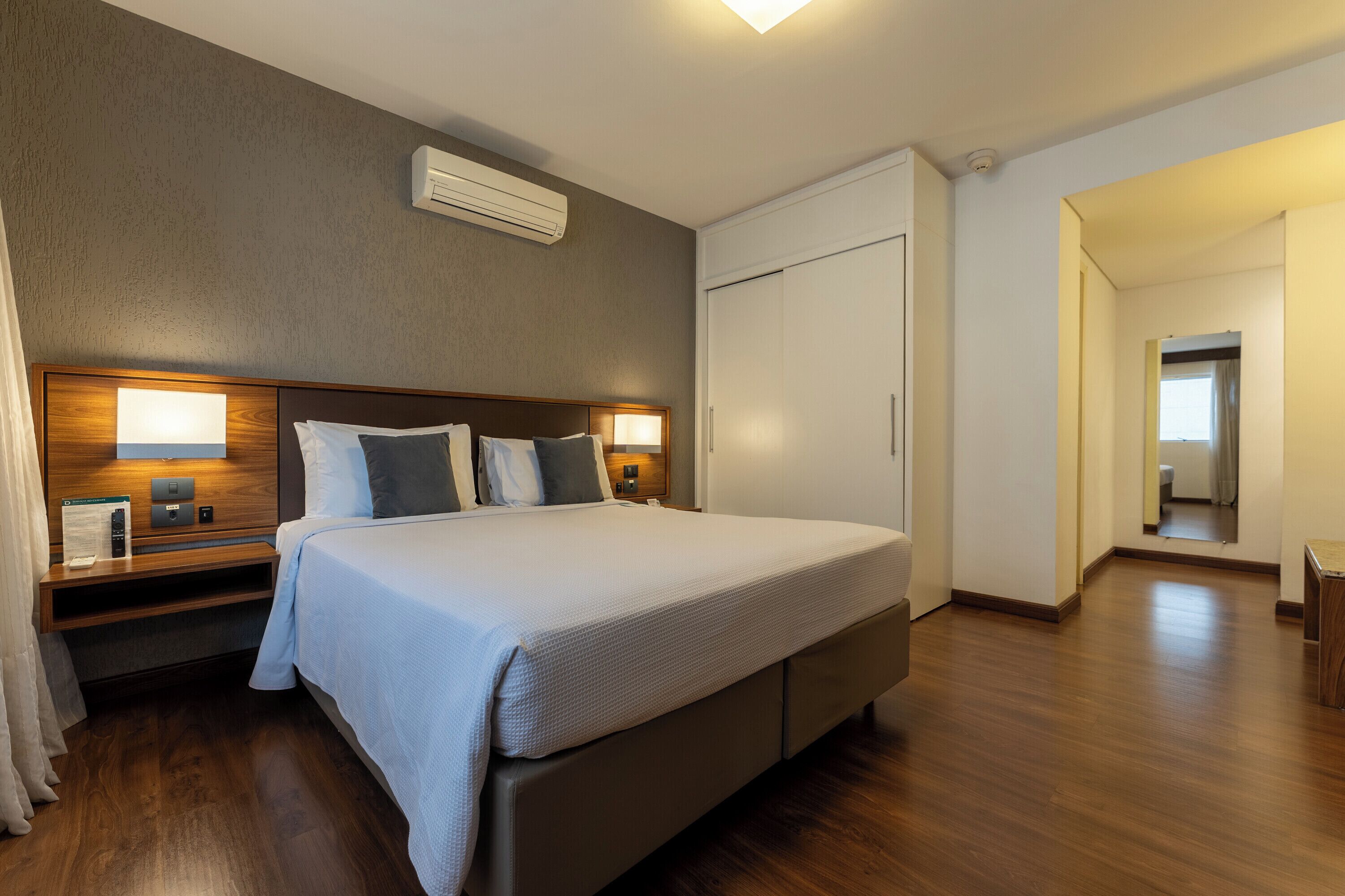 Quarto de hotel com cama grande, abajures, guarda-roupa e espelho.