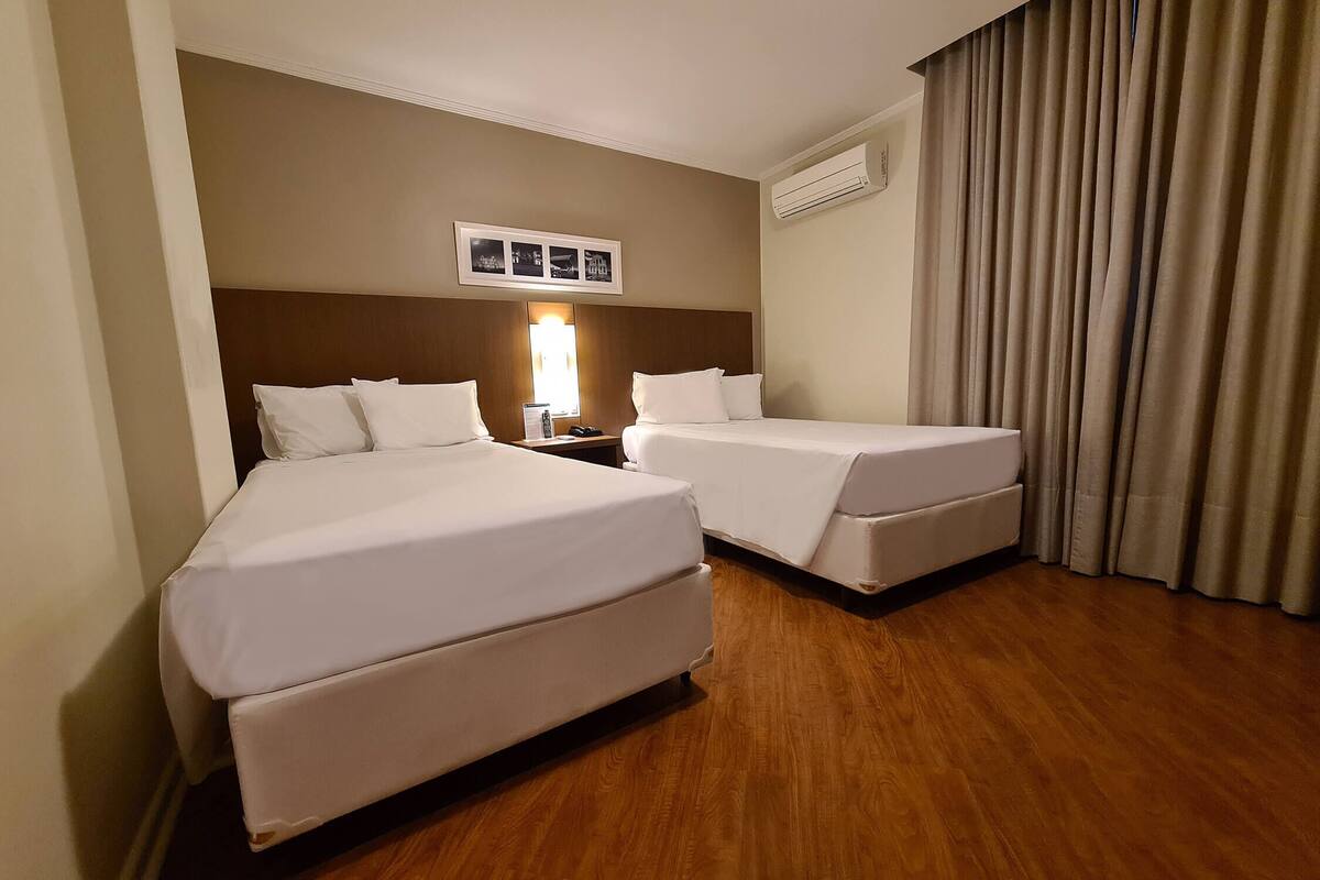 A hotel room with two beds, a nightstand with a lamp, a wall-mounted air conditioner, and a window with curtains.
