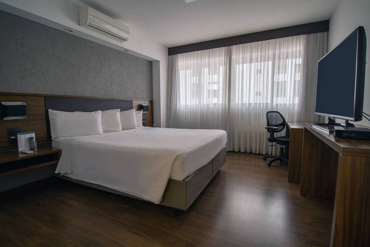 A hotel room with a large bed, a desk with a computer, a chair, and a window with curtains.