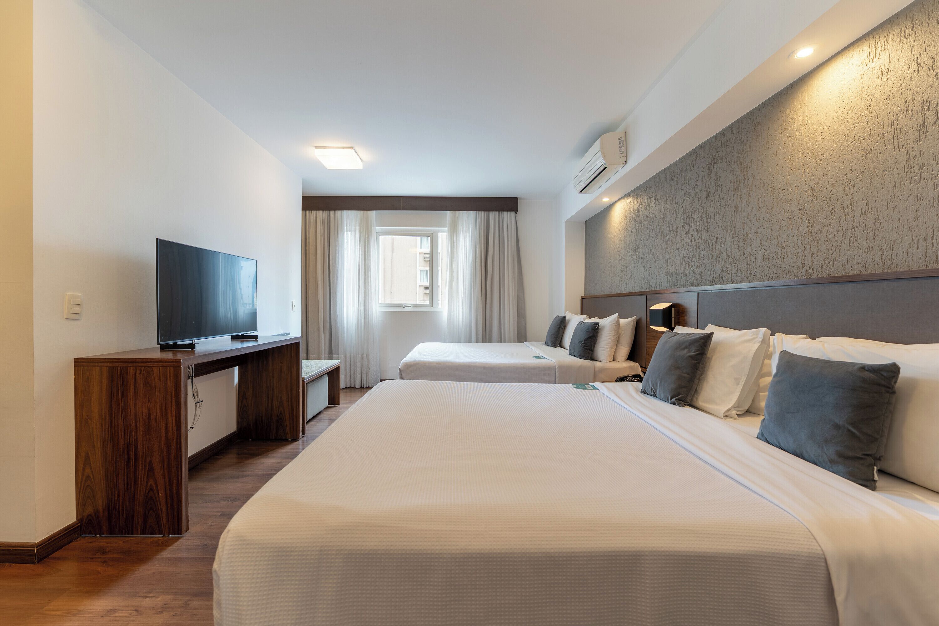 A hotel room with two beds, a desk with a flat-screen TV, a window with curtains, and an air conditioning unit on the wall.