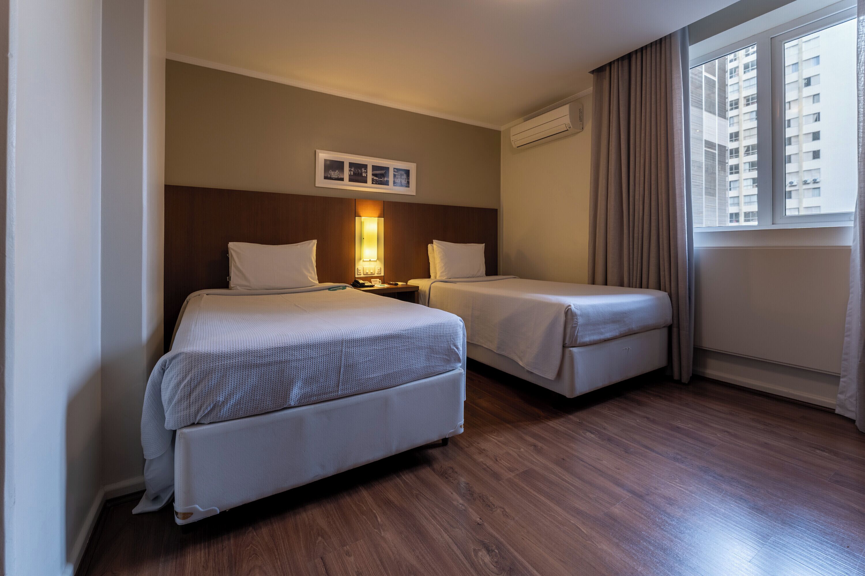A hotel room with two beds, a nightstand, a wall-mounted lamp, and a window with curtains.