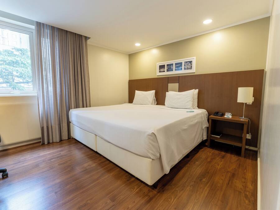 Habitación de hotel con una cama grande, un escritorio, una silla, una mesita de noche y un ventanal con cortinas.