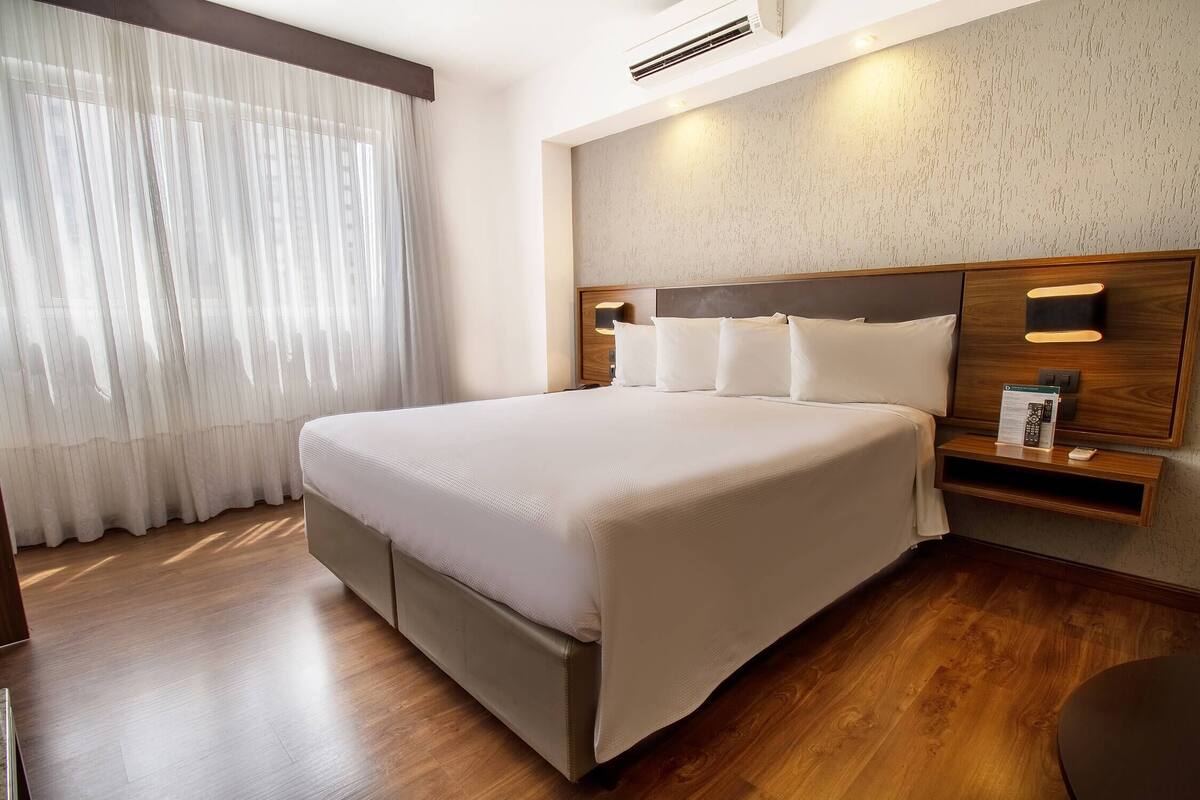 A hotel room with a large bed, a bedside table with a lamp, a wall-mounted shelf with a remote control, and a window with sheer curtains.