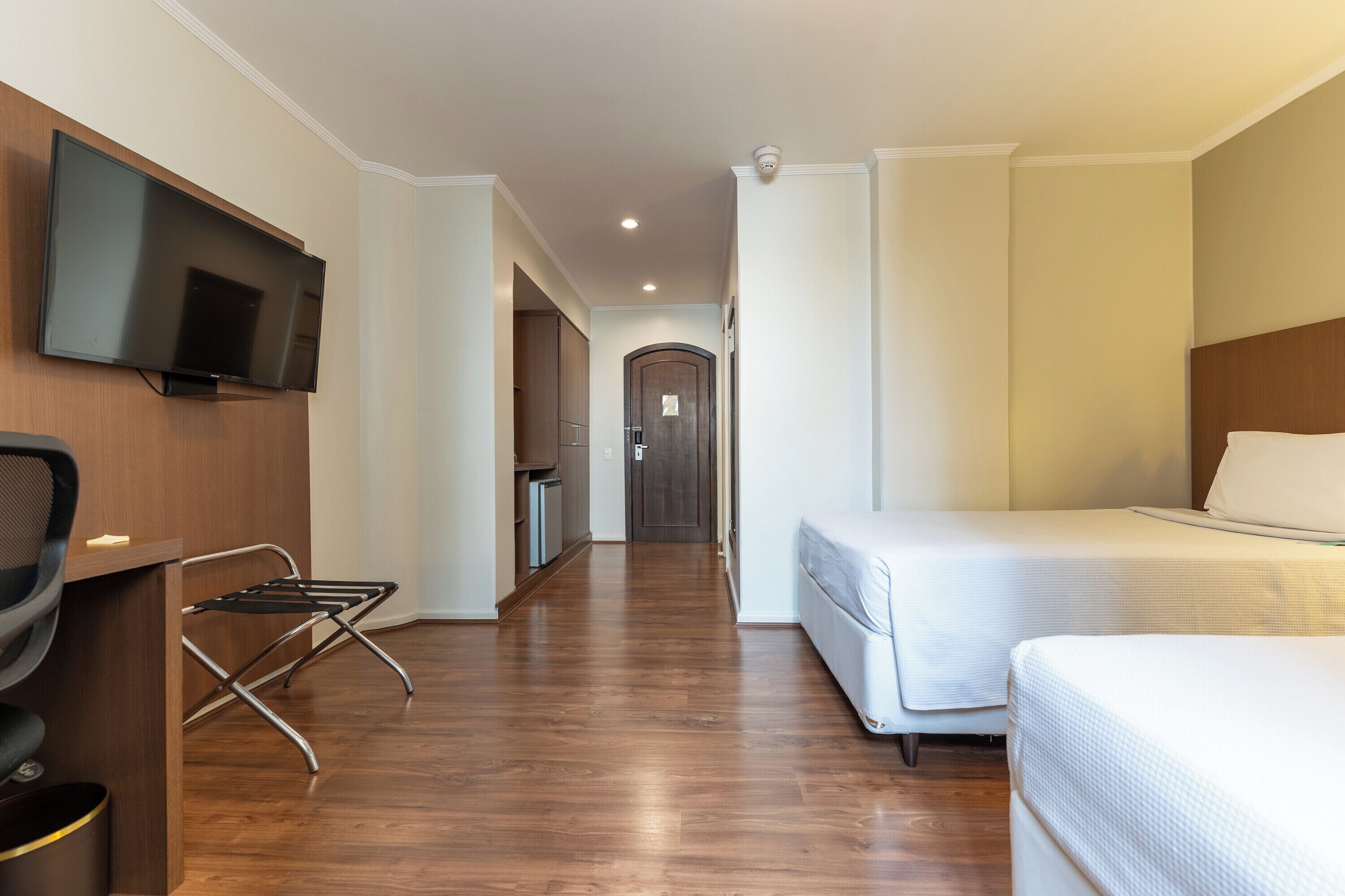 Habitación de hotel con cama, televisor montado en la pared, escritorio con silla y una kitchenette pequeña.