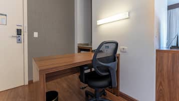 A hotel room with a wooden desk, a black office chair, a small trash bin, and a wall-mounted light fixture.