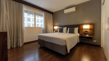 A hotel room with a large bed, two bedside tables with lamps, a window with curtains, and a wall-mounted air conditioner.