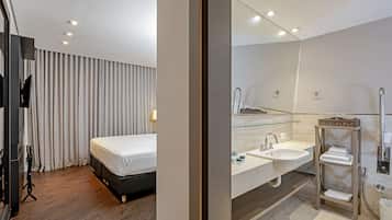A modern hotel room with a bed, a large mirror, a vanity with a sink, and a shower area.