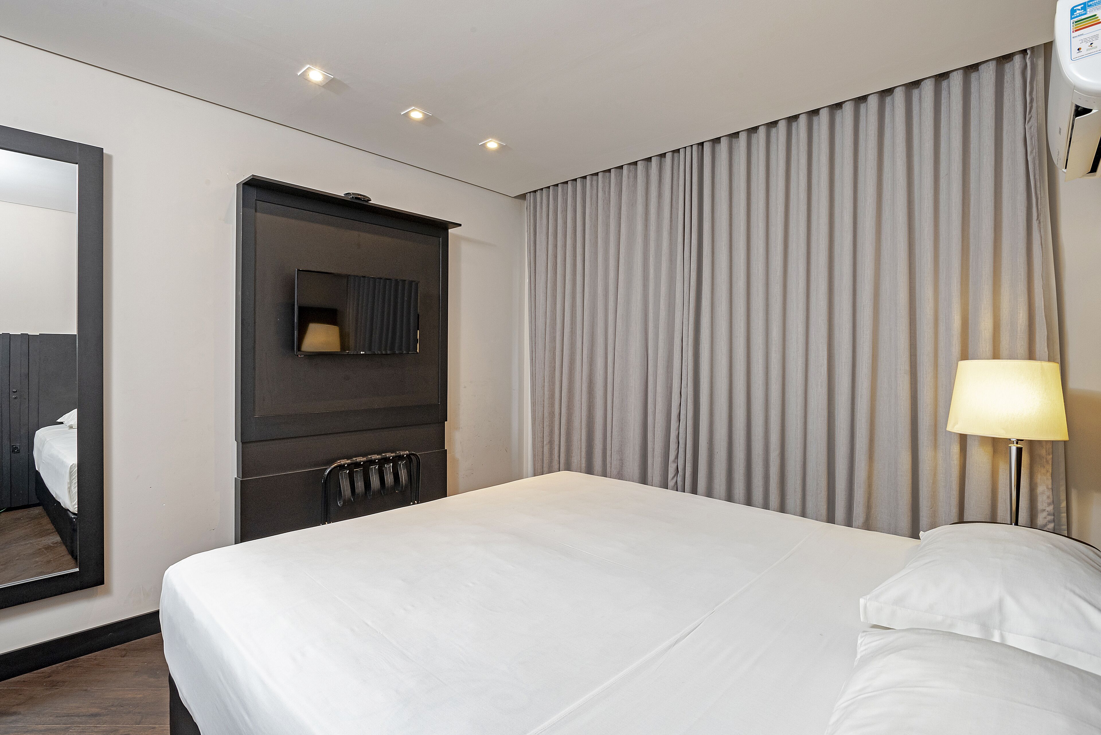 A hotel room with a large bed, a dark headboard, a mirror, a wall-mounted TV, and a window with curtains.