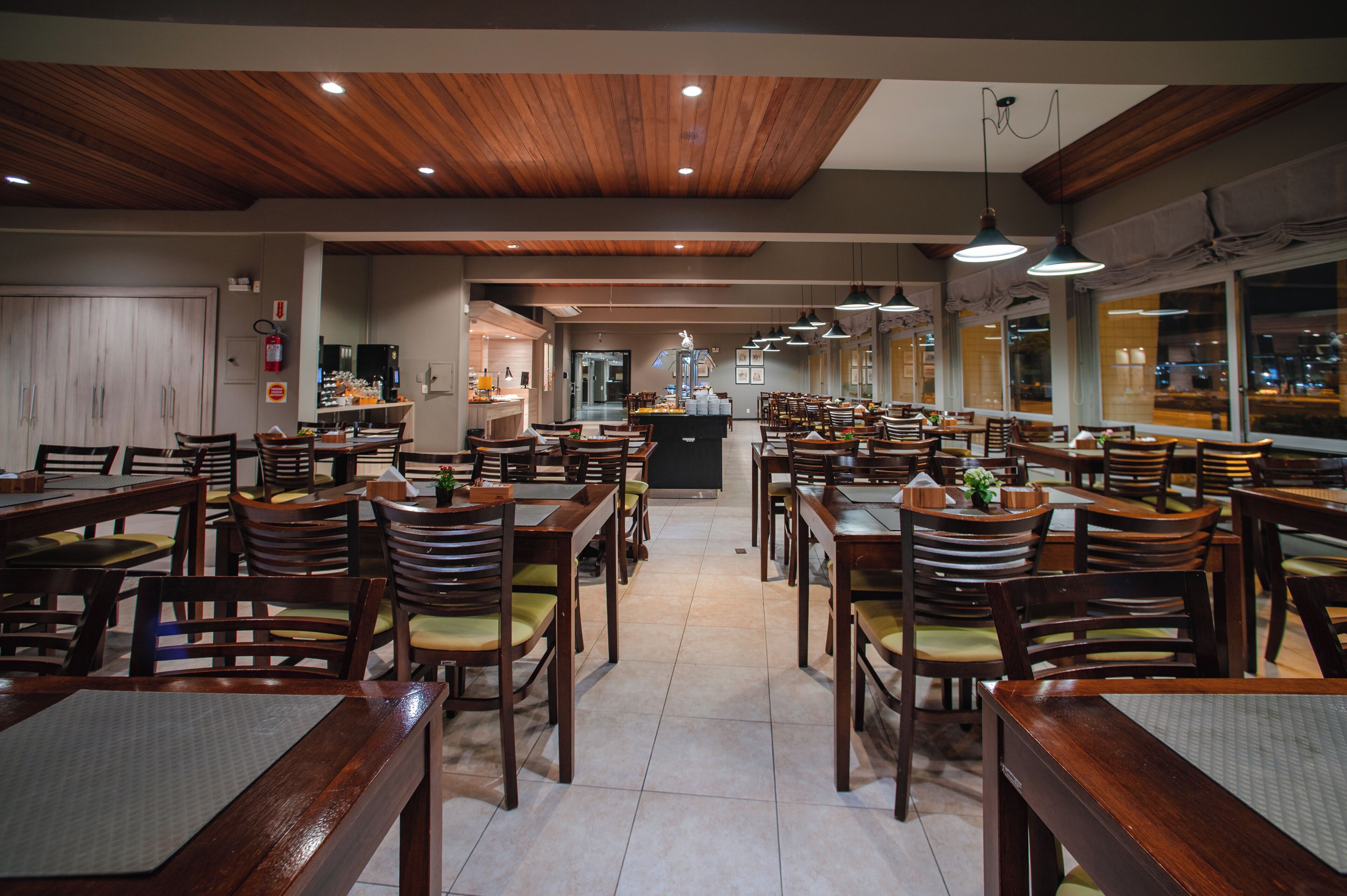 A spacious restaurant with wooden tables and chairs, a kitchen area in the background, and large windows.