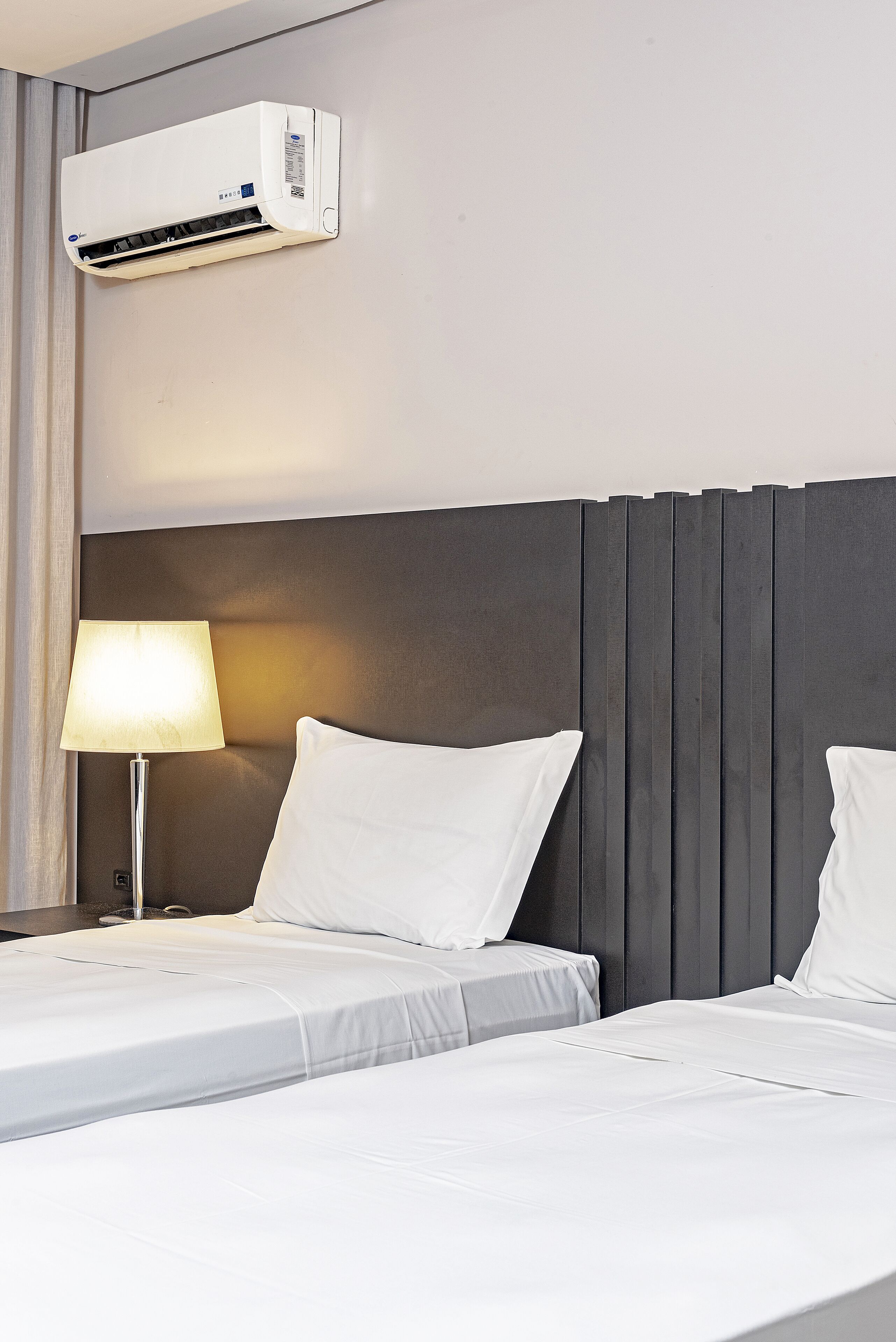 Habitación de hotel con dos camas, aire acondicionado de pared, lámpara de noche y cabecera oscura.