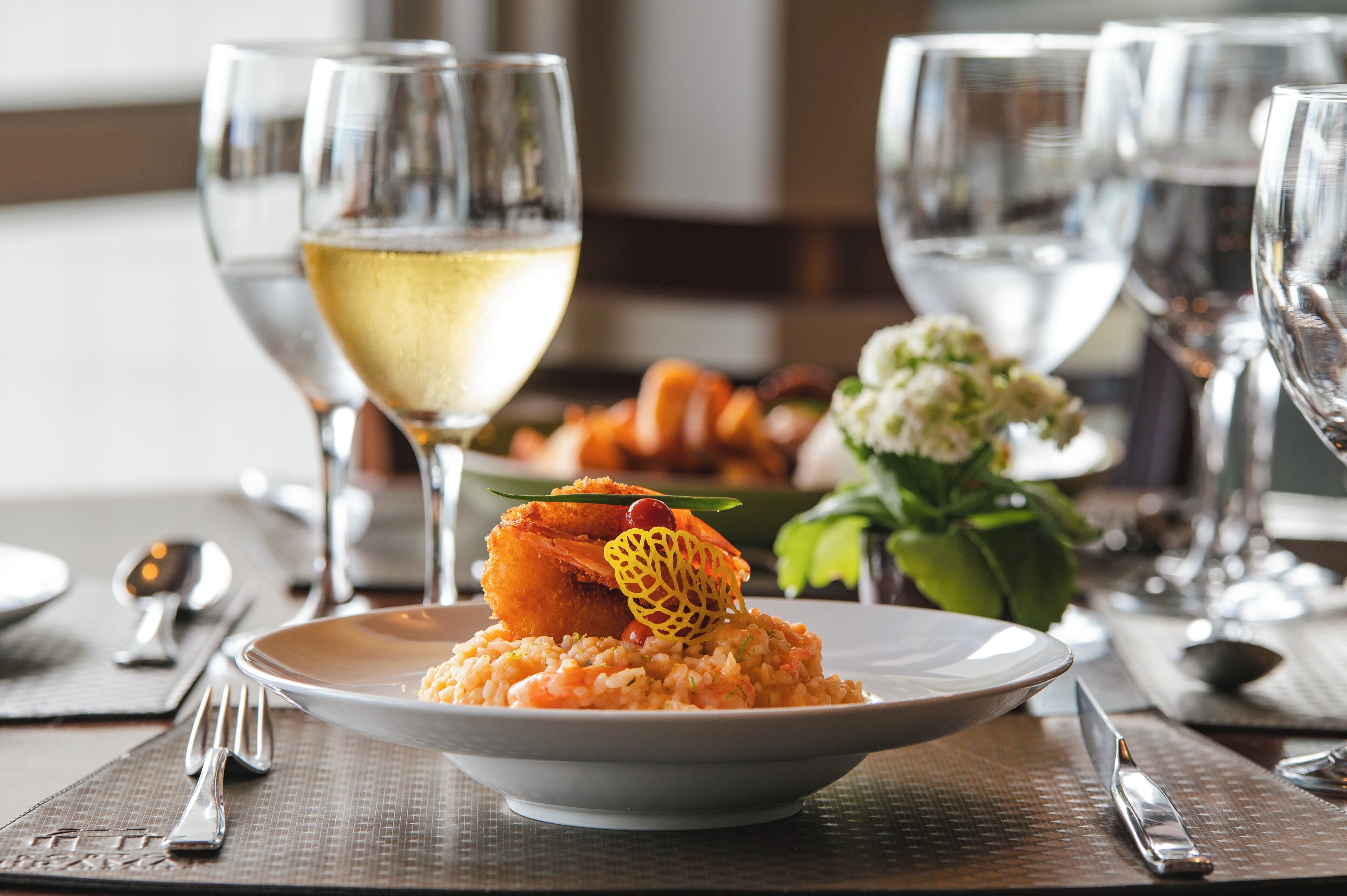 Una mesa de comedor con un plato de comida, una copa de vino blanco y un florero con flores.