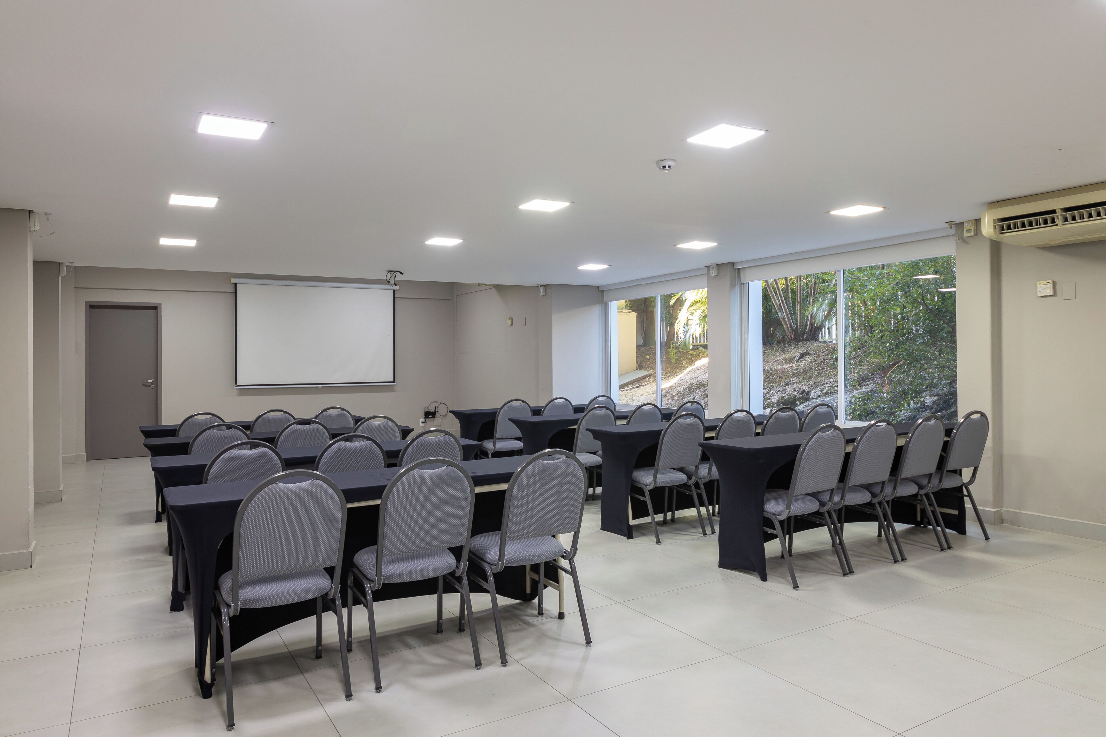 Un salón de conferencias con mesas, sillas, una pantalla de proyector y ventanas amplias.