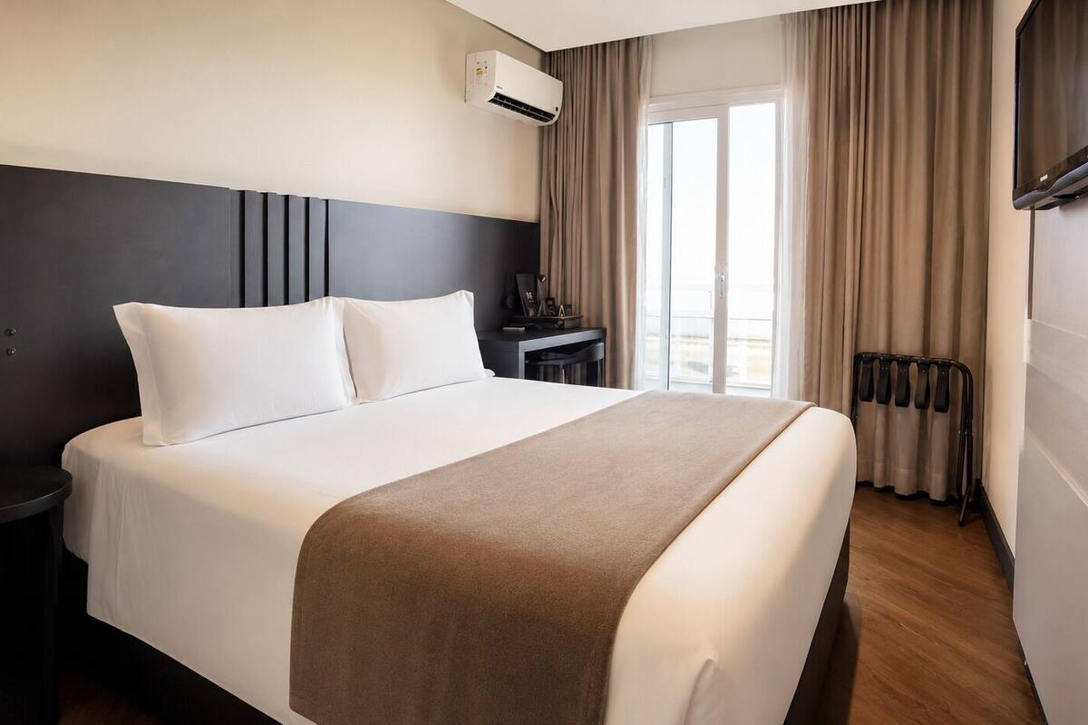 Ett hotellrum med en säng, ett sängbord, ett skrivbord, en stol, en luftkonditioneringsanläggning, ett element och ett fönster med gardiner.