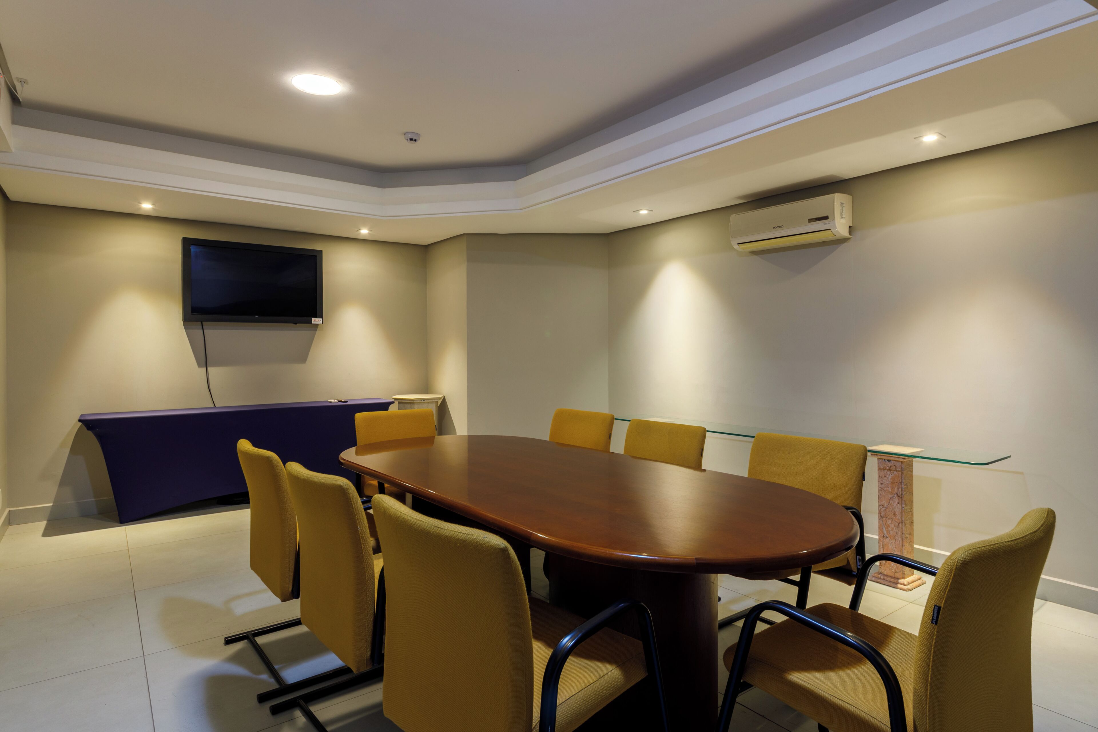 A conference room with a long table, yellow chairs, a flat-screen TV, and an air conditioning unit.