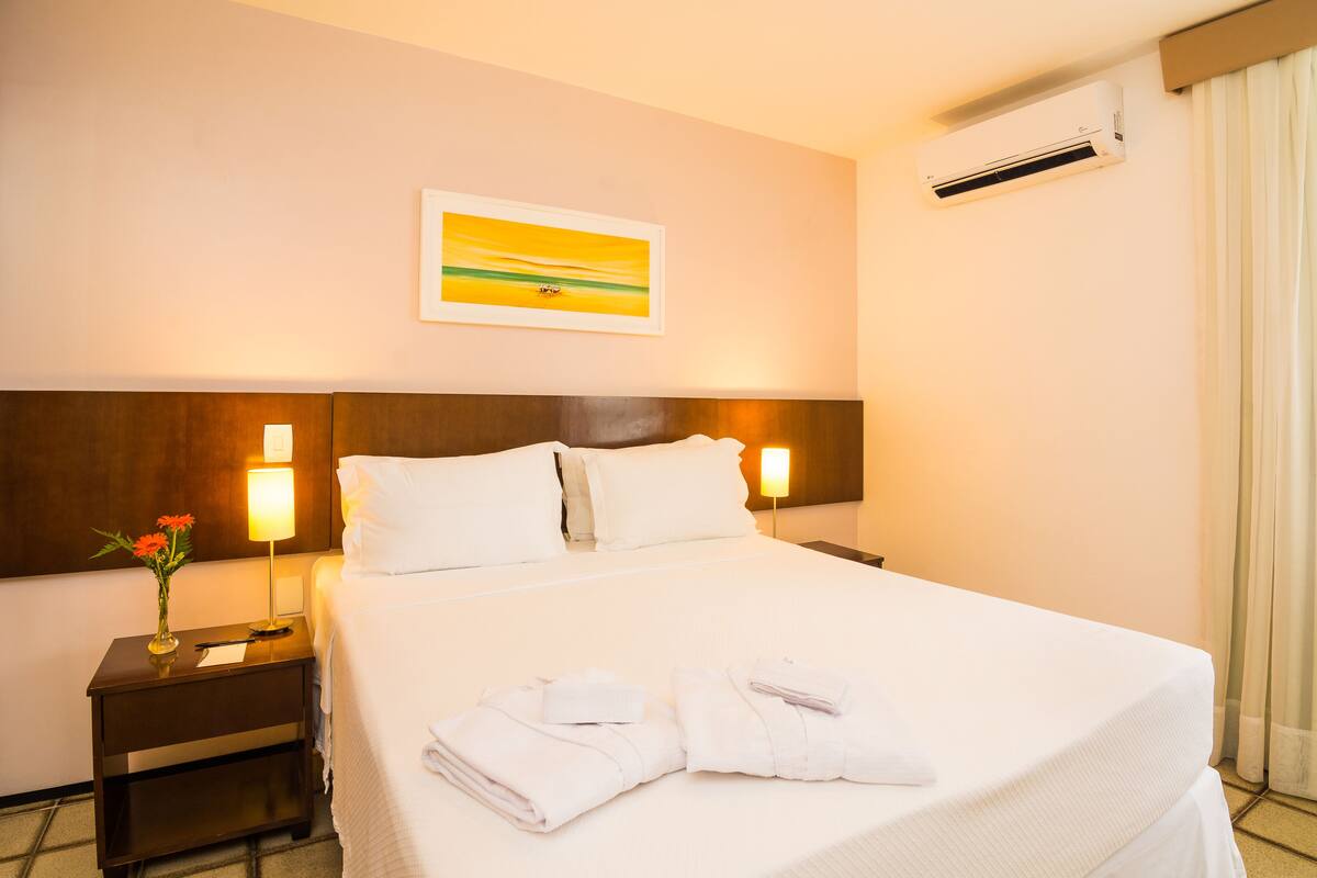 A hotel room with a neatly made bed, two bedside lamps, a framed picture on the wall, and an air conditioning unit.