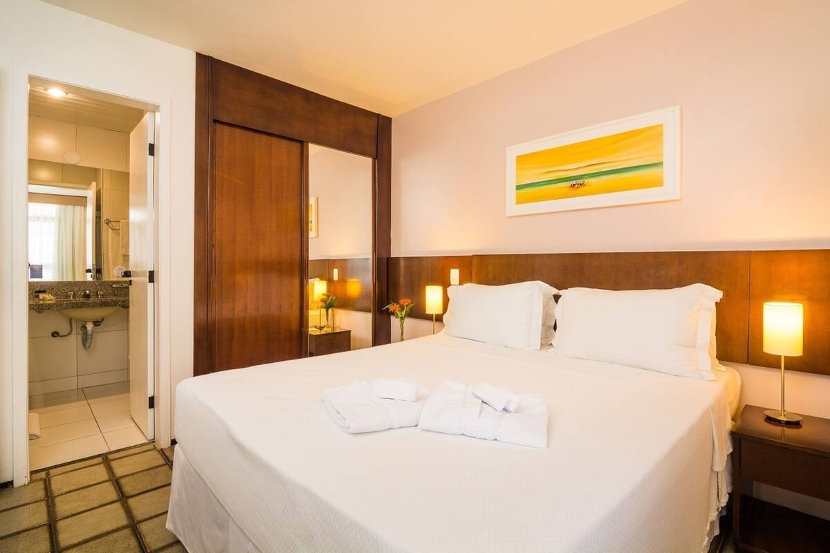 A hotel room with a large bed, a wooden headboard, a painting on the wall, a bedside lamp, and a bathroom visible through an open door.