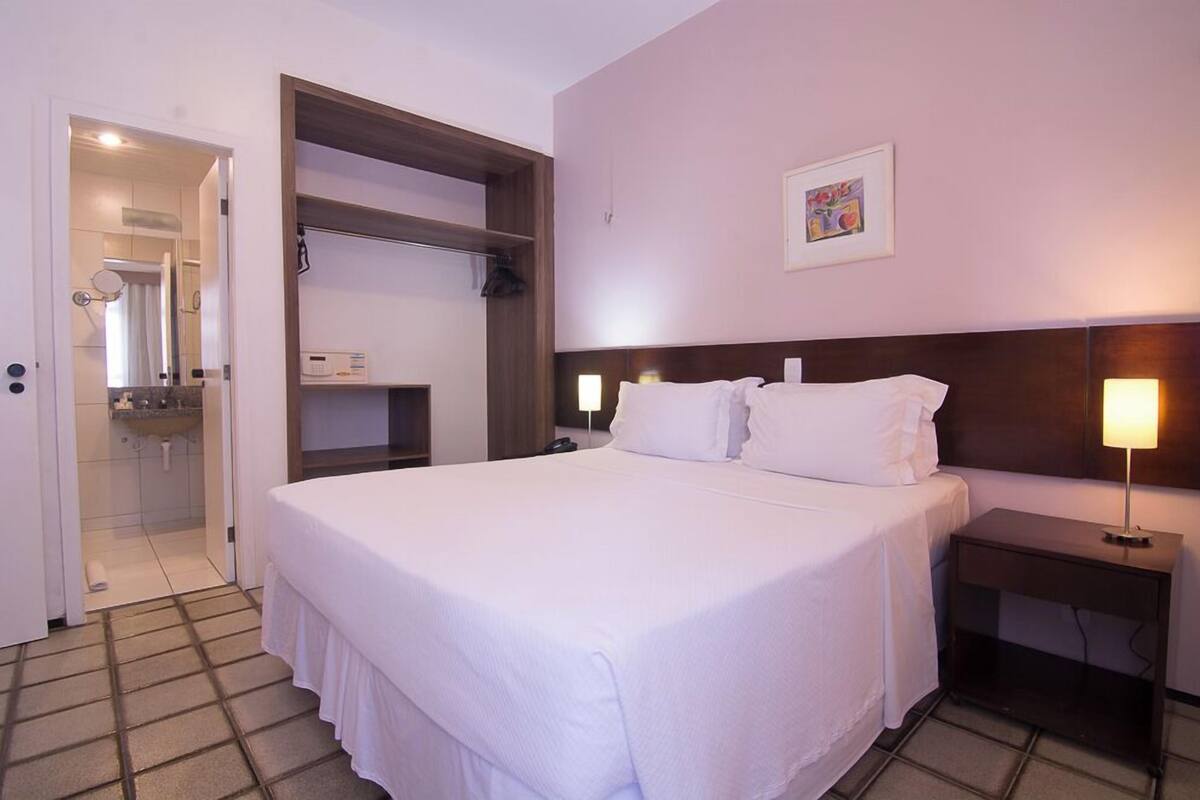 A hotel room with a large bed, a bedside table with a lamp, a wardrobe, and a shower area.